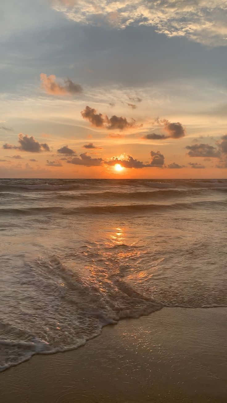 晴空日落海灘圖 壁紙 — 金色夕陽照耀在帶有零星雲朵的沙灘上翻滾的海浪上。