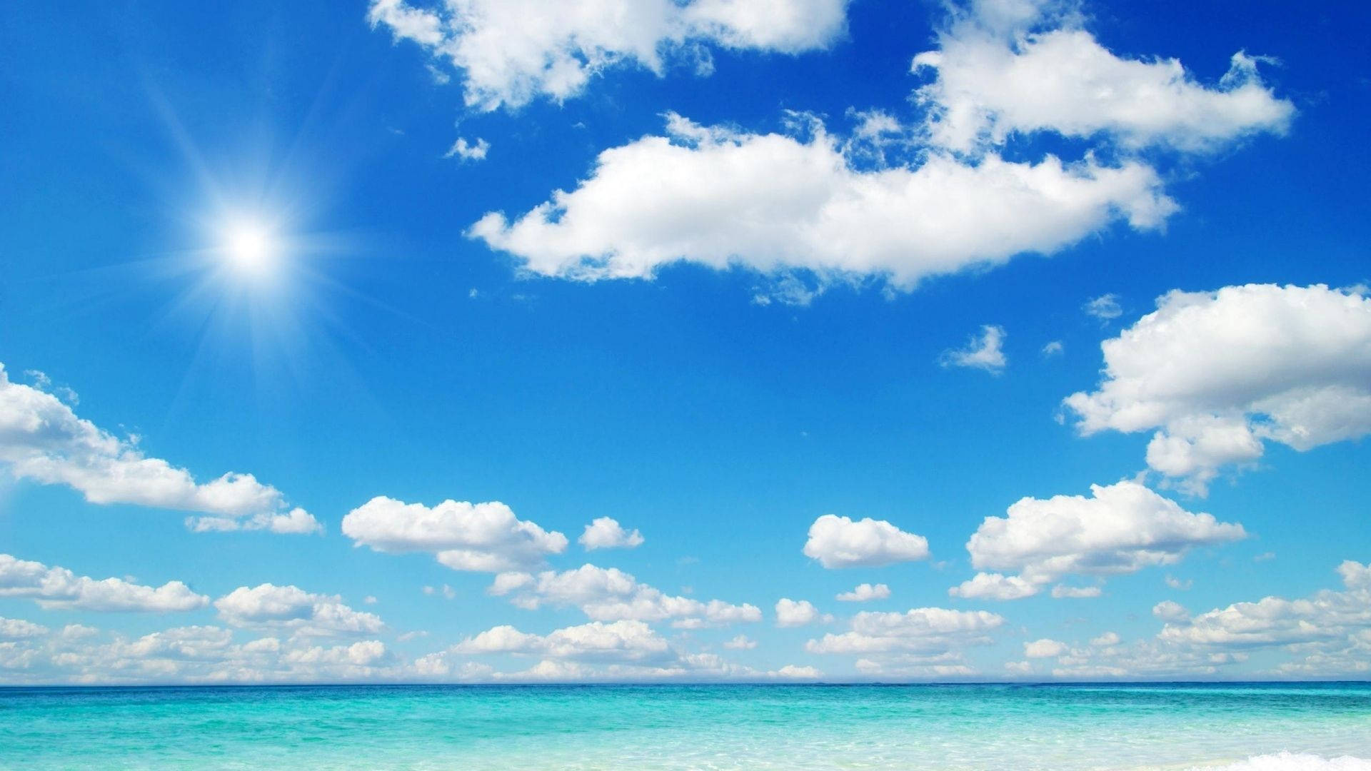 晴空海景 壁紙 — 充滿活力的綠松石海洋與燦爛的藍天相遇，天空中有蓬鬆的白雲和明亮的太陽。