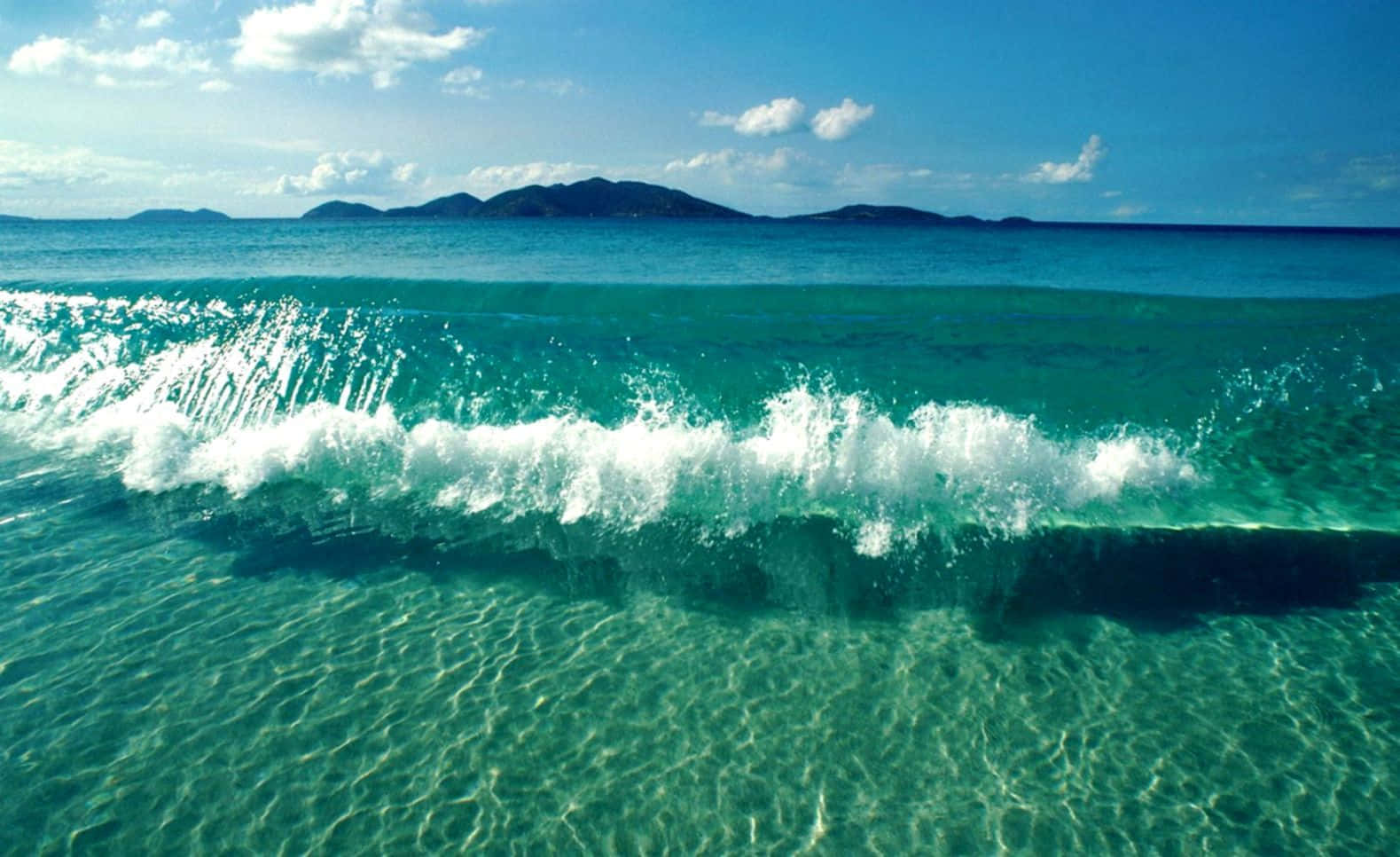 藍綠色的海浪在沙底附近帶著白色泡沫翻騰，遠處有島嶼。