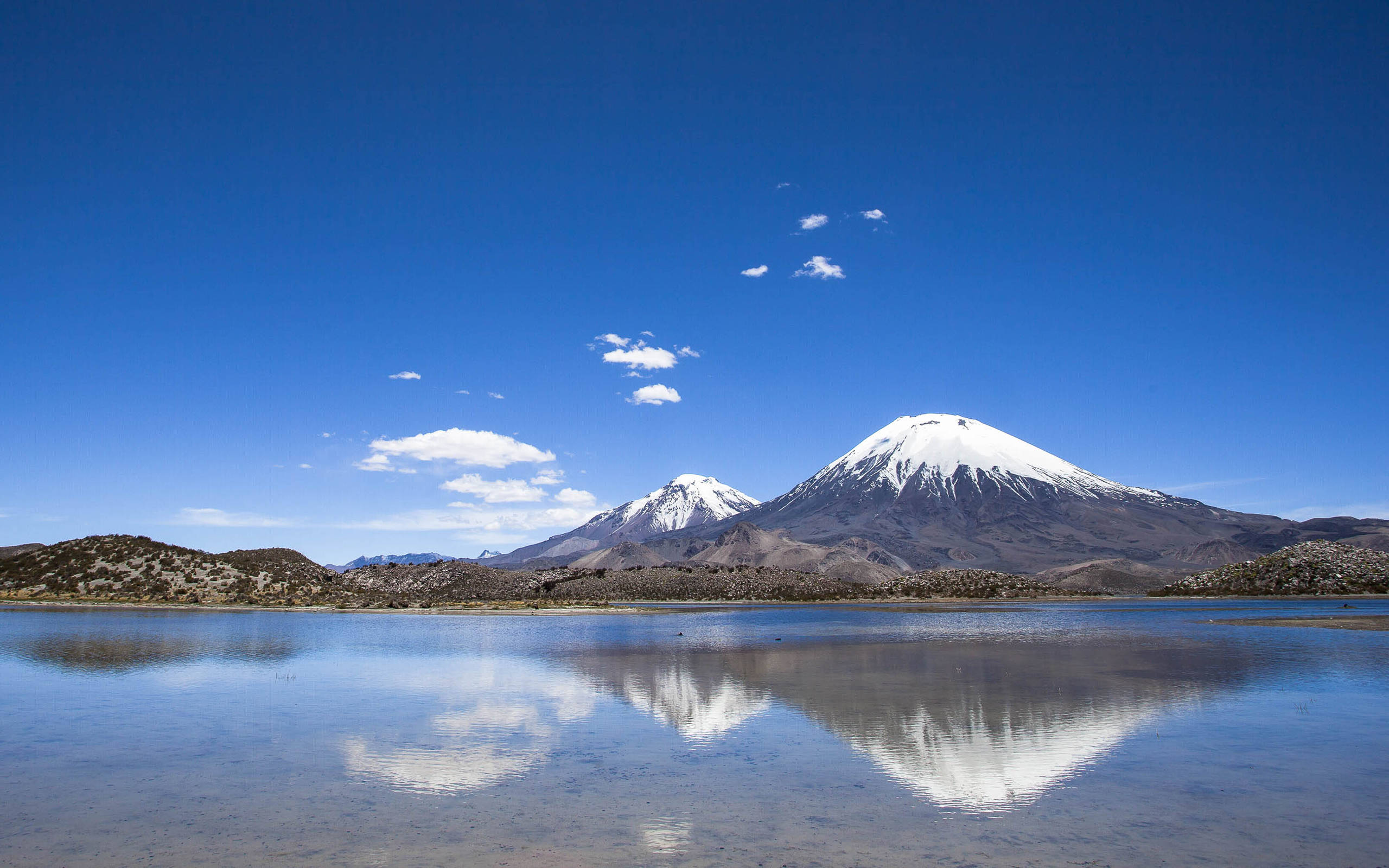 智利勞卡國家公園壯麗景色 壁紙 — 白雪覆蓋的火山在平靜的湖泊後方雄偉地聳立，在清澈藍天與零星雲朵下倒映著它們的山峰。
