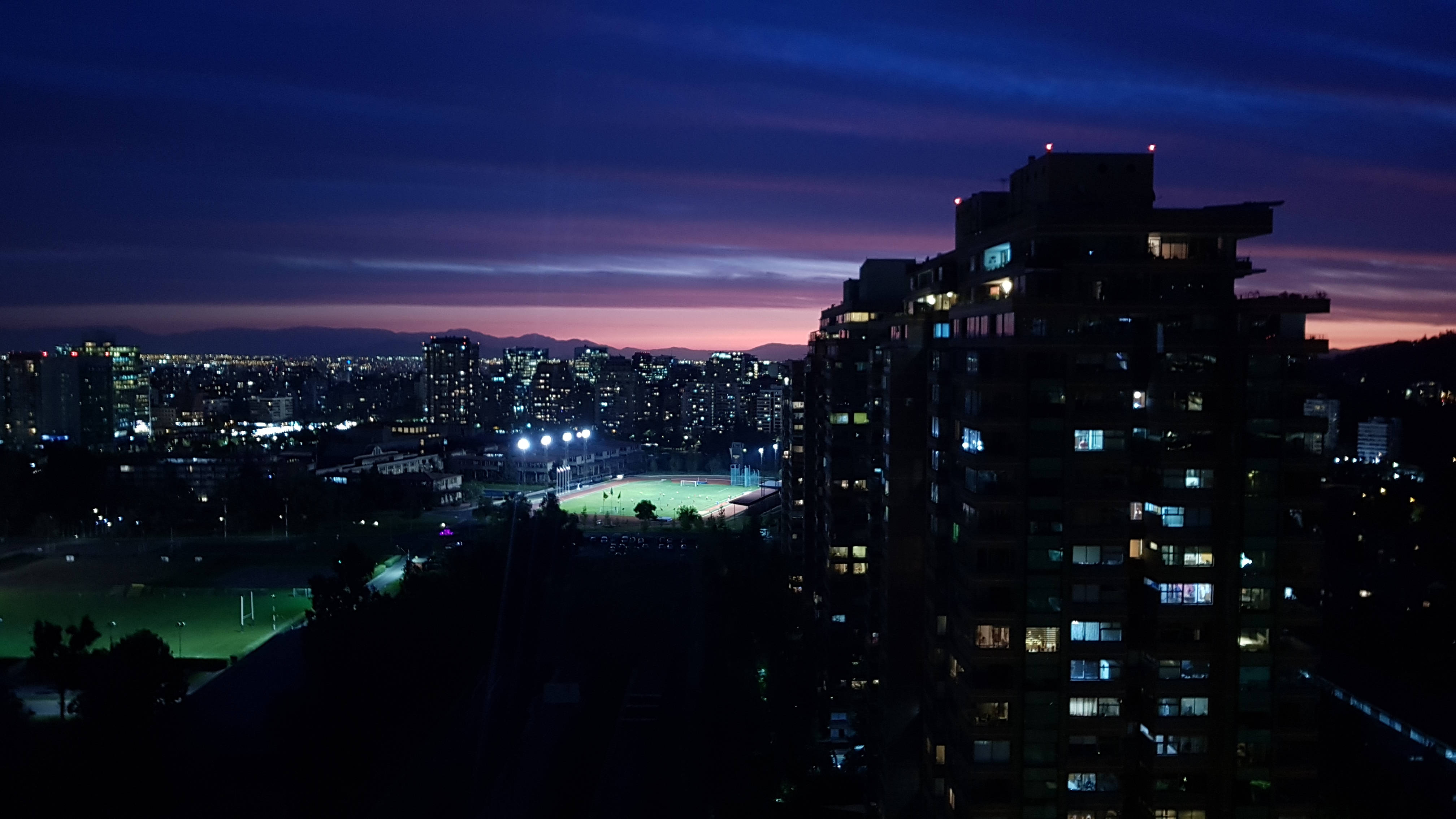智利聖地亞哥夜幕初降 壁紙 — 黃昏時分的城市景觀，在紫色和藍色的天空下，有燈火通明的高層建築和下方亮燈的運動場。