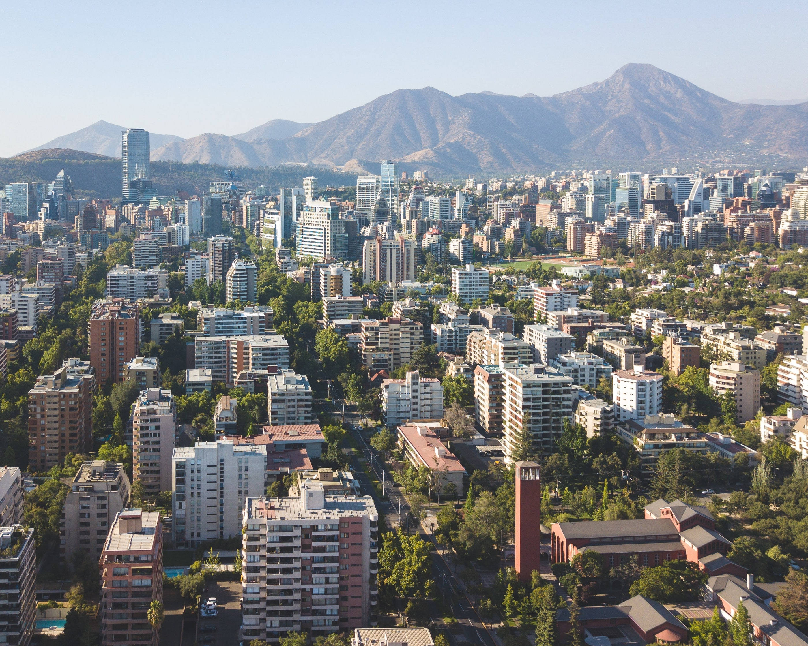 智利聖地亞哥山景 壁紙 — 從高空俯瞰一座廣闊的城市，現代建築與茂密綠植交織，背景是雄偉的山脈，天空湛藍清澈。