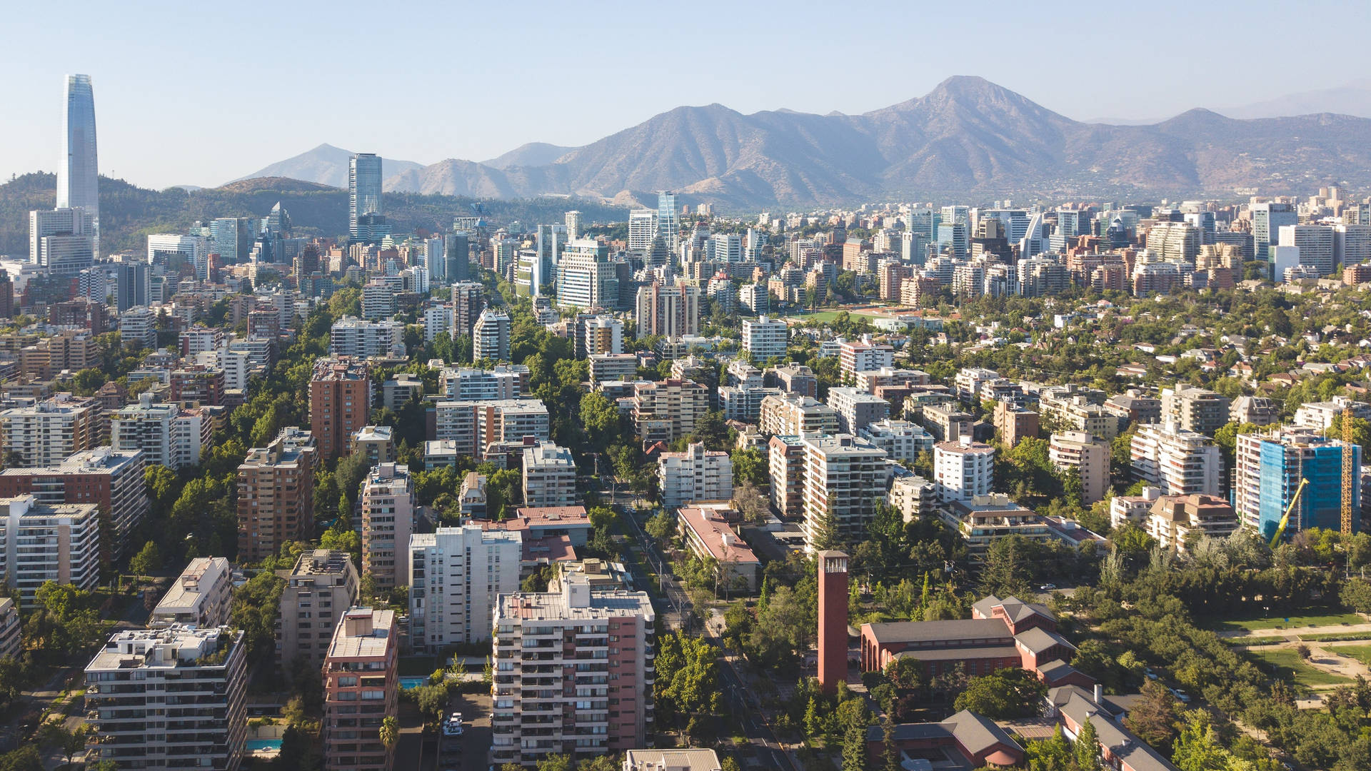 智利聖地亞哥空中俯瞰 壁紙 — 鳥瞰一個廣闊的城市，現代摩天大樓和綠色公園依偎在崎嶇山脈的背景下，天空清澈湛藍。