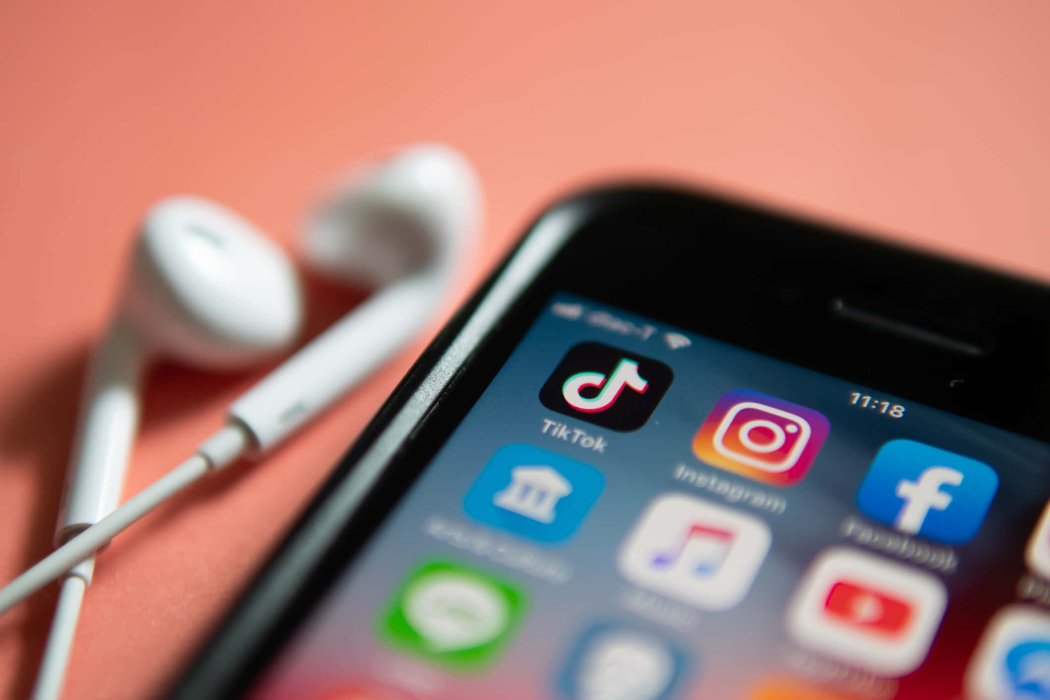 Social Media 桌布 壁紙 — 智能手機特寫，顯示社交媒體應用程式，白色耳機放在旁邊的珊瑚色背景上。