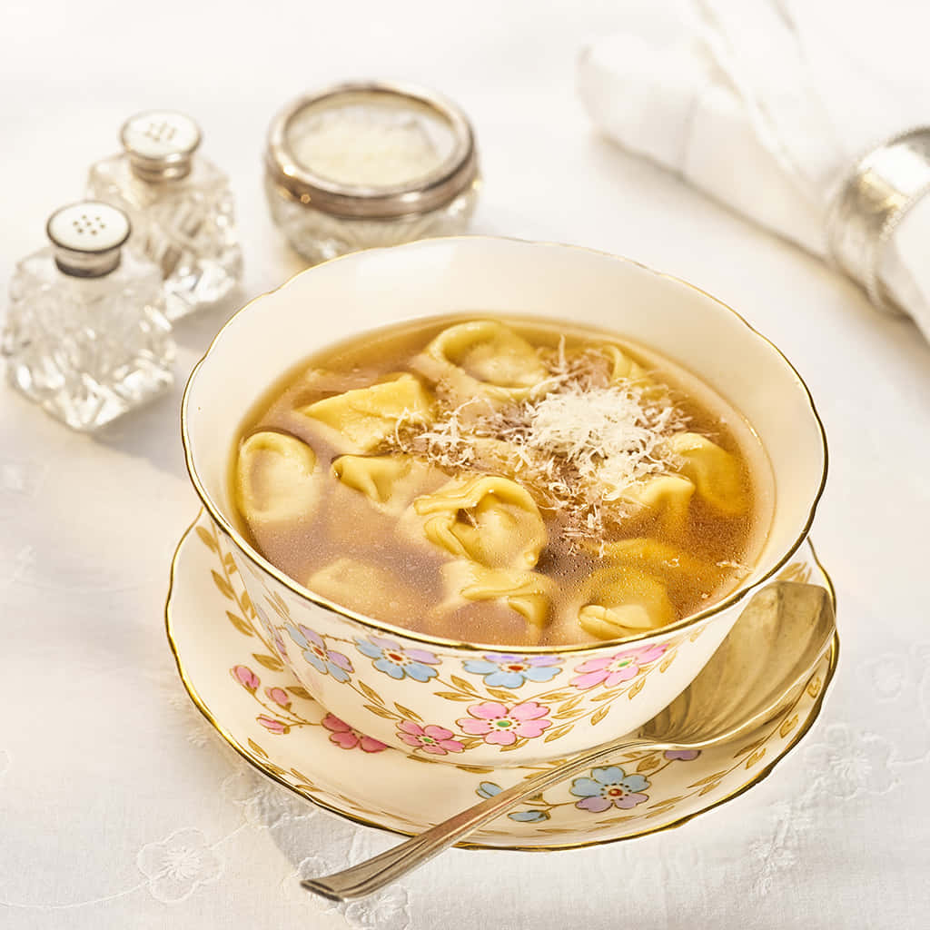 暖香濃郁的意大利餃子湯 壁紙 — 一碗冒煙的意大利餃子湯，撒上磨碎的芝士，優雅地放在花卉圖案的碟子上，旁邊有水晶鹽和胡椒瓶。
