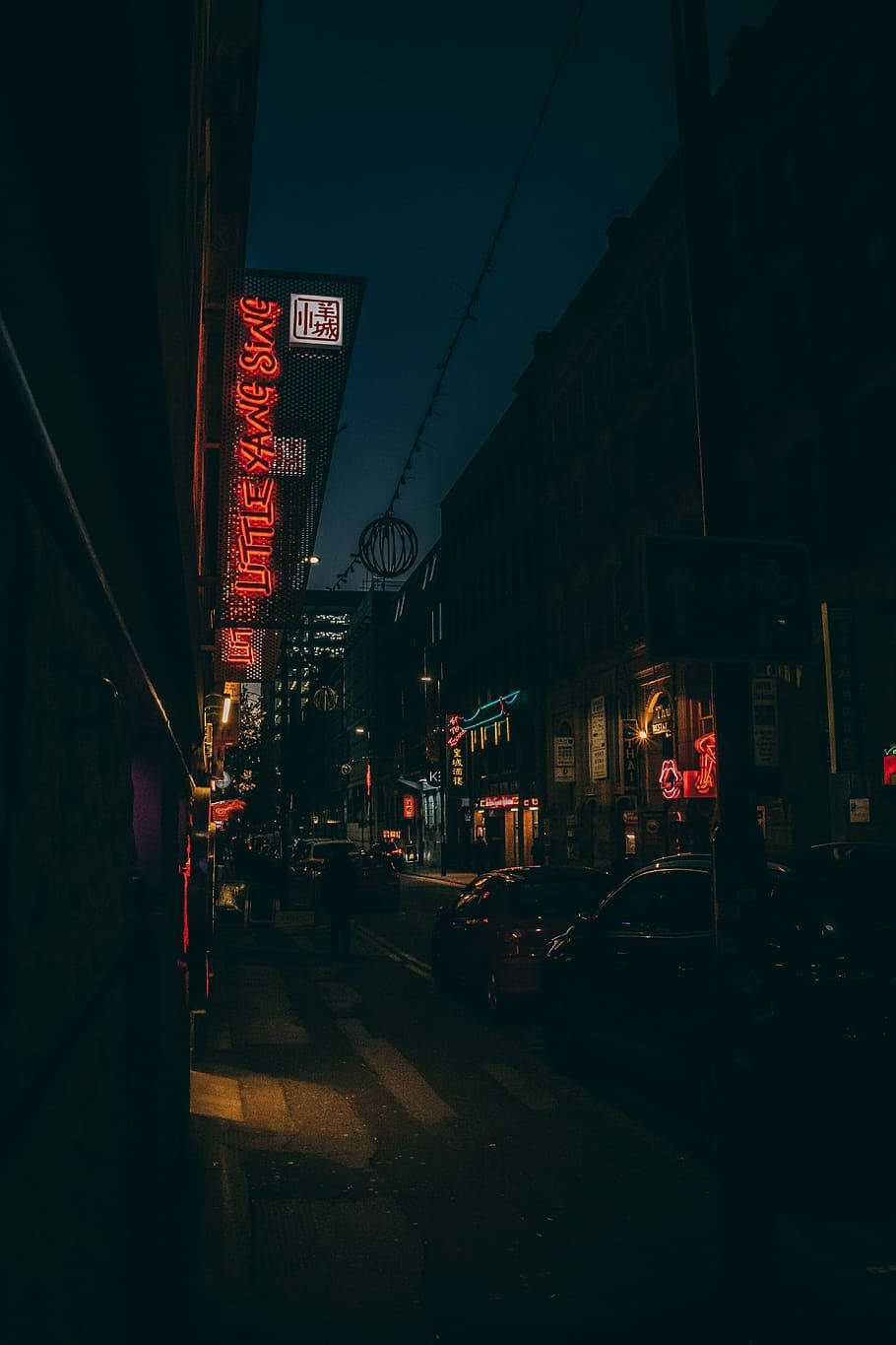暗城霓虹街燈 壁紙 — 一條狹窄的城市街道在夜晚被發光的紅色霓虹燈招牌照亮，在潮濕的路面上投射出溫暖的光芒。