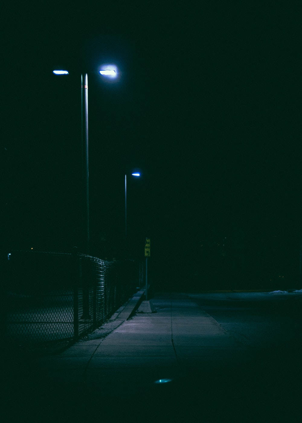 夜晚一條寧靜空無一人嘅行人路，被藍色街燈照亮，旁邊有鐵絲網圍欄。