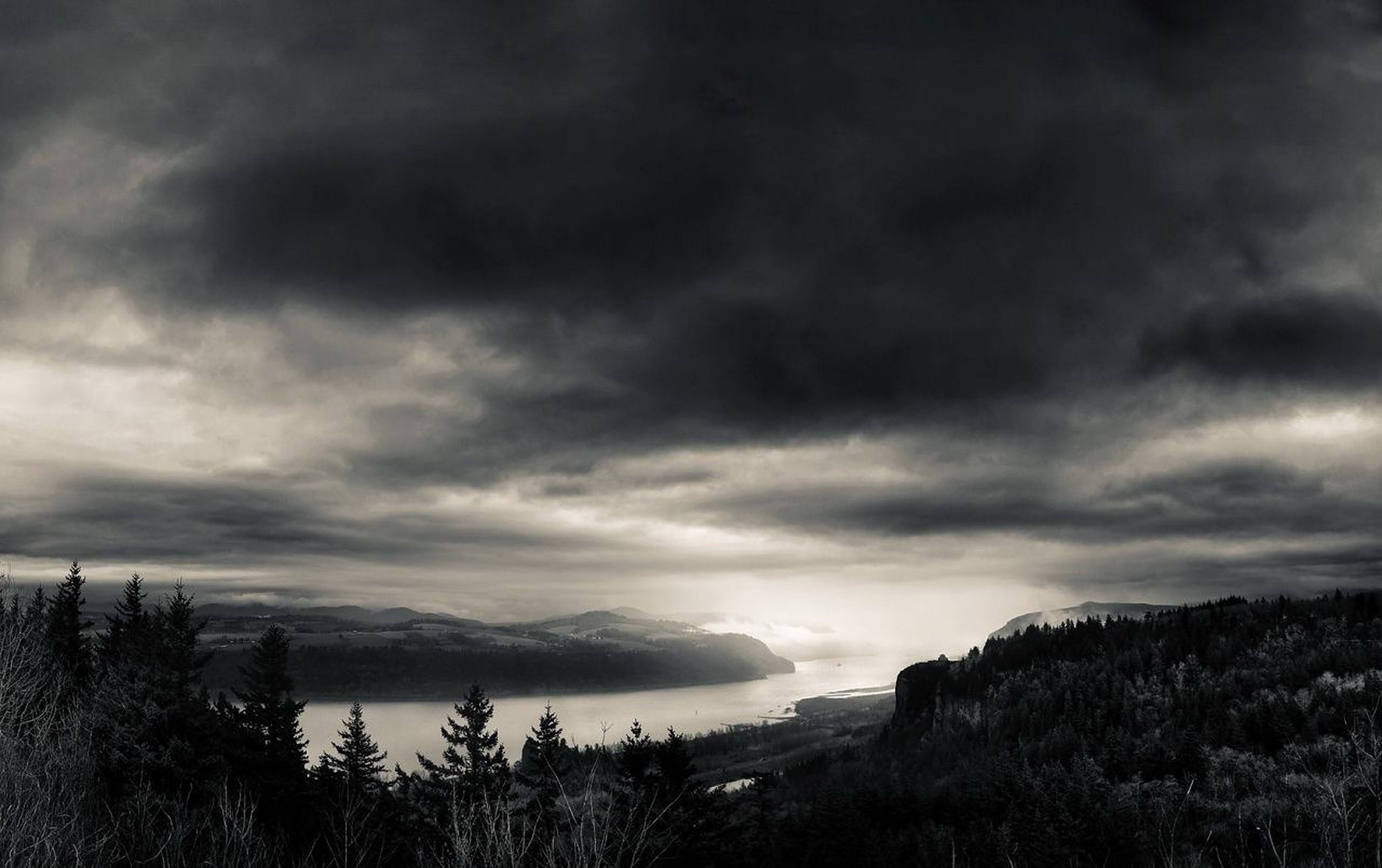 暗夜天氣 壁紙 — 一幅陰鬱的黑白風景畫，展示了一個霧氣瀰漫的湖泊，坐落在森林覆蓋的山丘之間，天空烏雲密布。