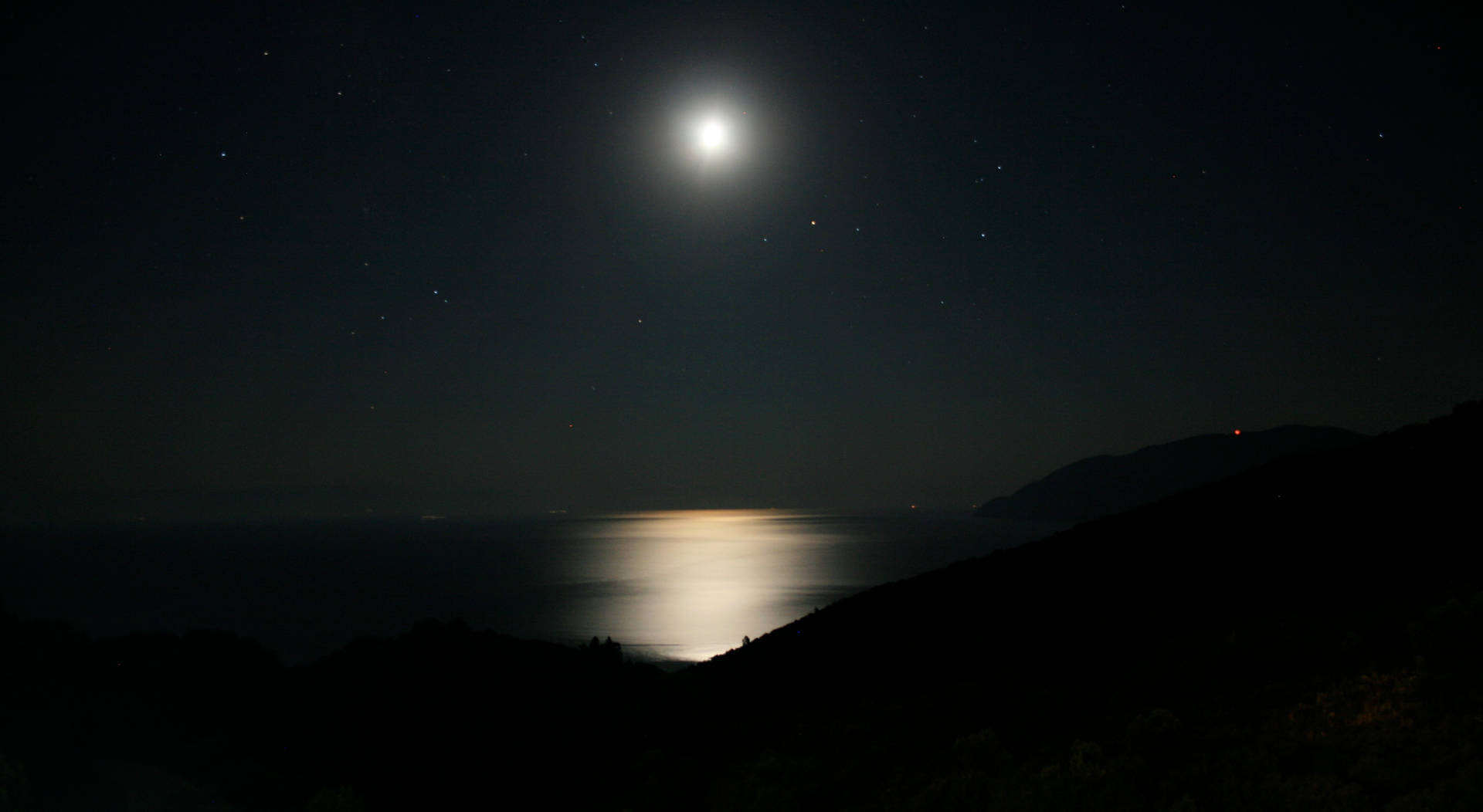 月光在夜間反射在海面上，星空和黑暗的山丘。