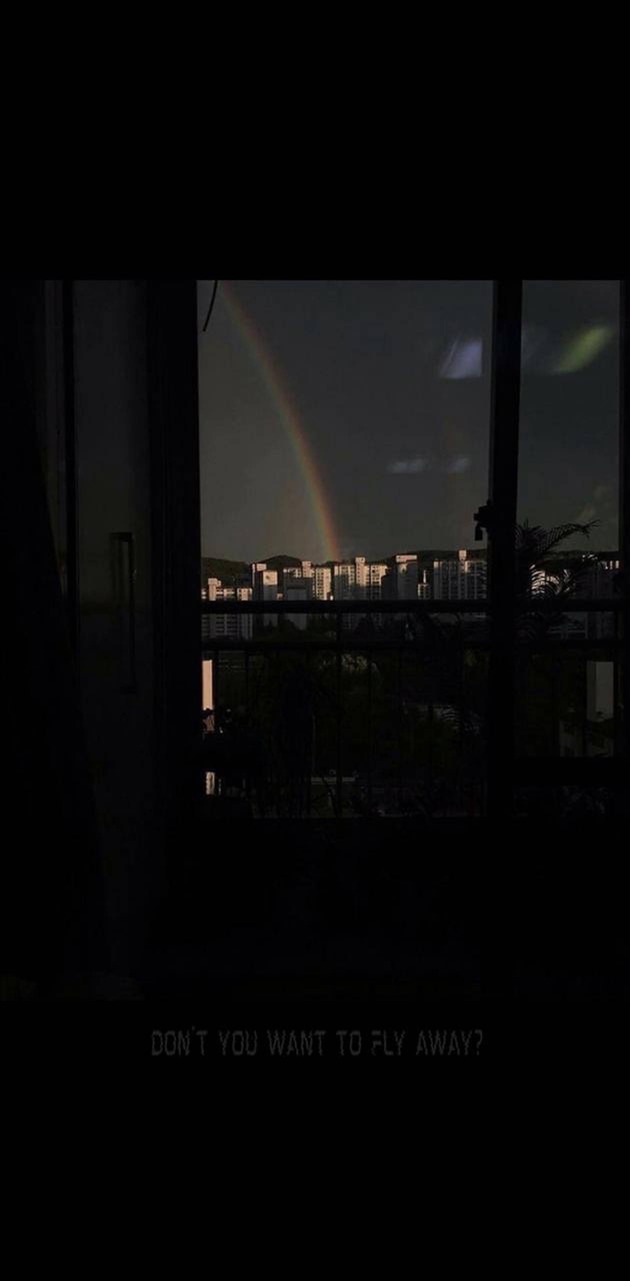 暗夜都市中的彩虹 壁紙 — 一道微弱的彩虹拱越城市天際線，透過夜晚的暗窗觀看，建築燈光閃爍。