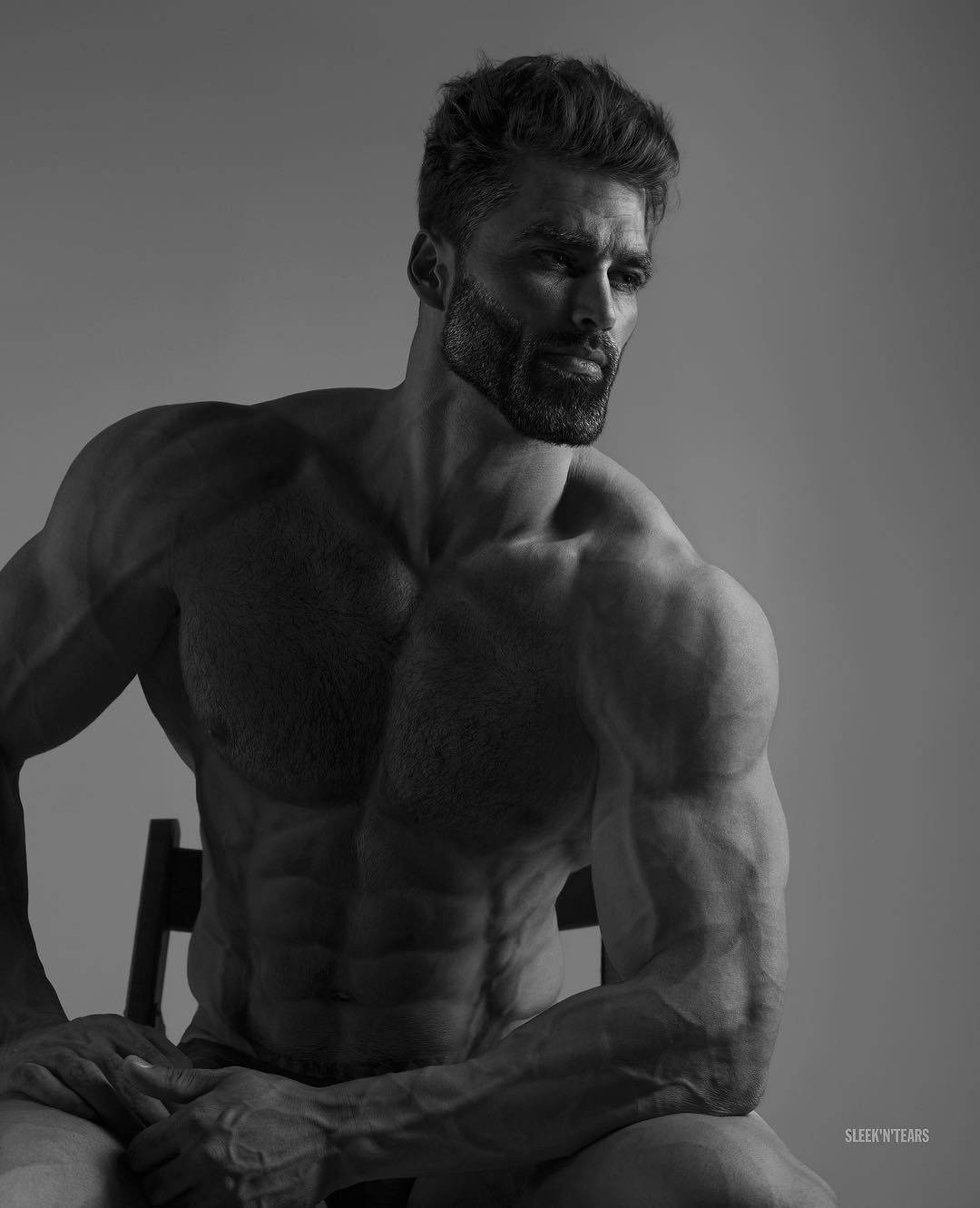 暗影中的Giga Chad 雄偉肖像 壁紙 — 一個留鬍鬚的肌肉男赤裸上身坐在椅子上，他分明的體格在戲劇性的黑白燈光下凸顯出來。