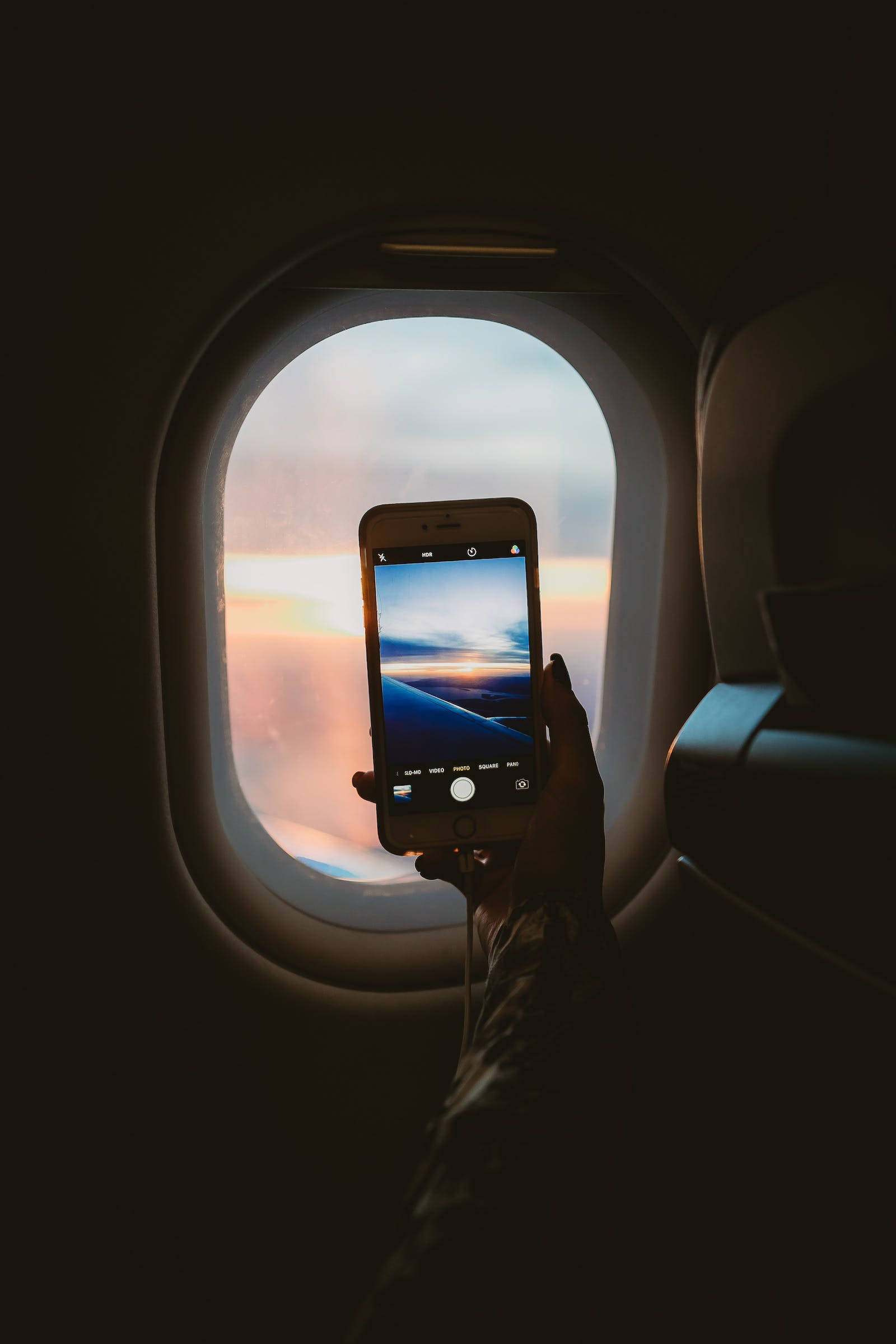 暗色iPhone飛機上拍攝影像 壁紙 — 一隻手拿著手機，透過飛機窗戶捕捉充滿活力的日落，伴隨著柔軟的雲朵和溫暖的色調。