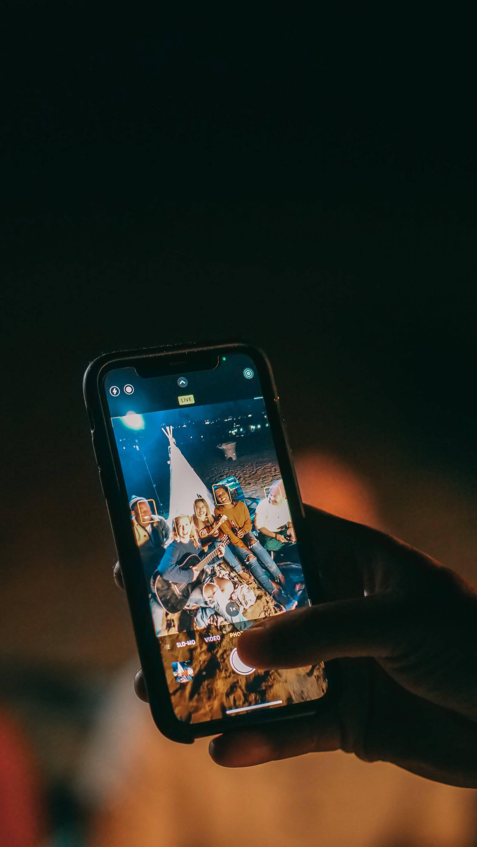 暗色iPhone飛機上拍攝影像 壁紙 — 一隻手拿著手機，顯示一張鮮豔的照片，照片中朋友們在星空下圍著營火，背景有一個帳篷。