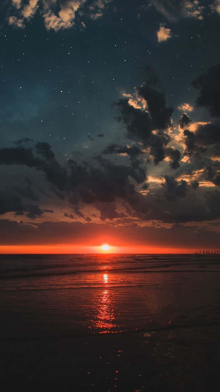 暗色天空的日落海灘圖 壁紙 — 海洋上空熾熱的日落，星星在戲劇性的多雲天空中出現。