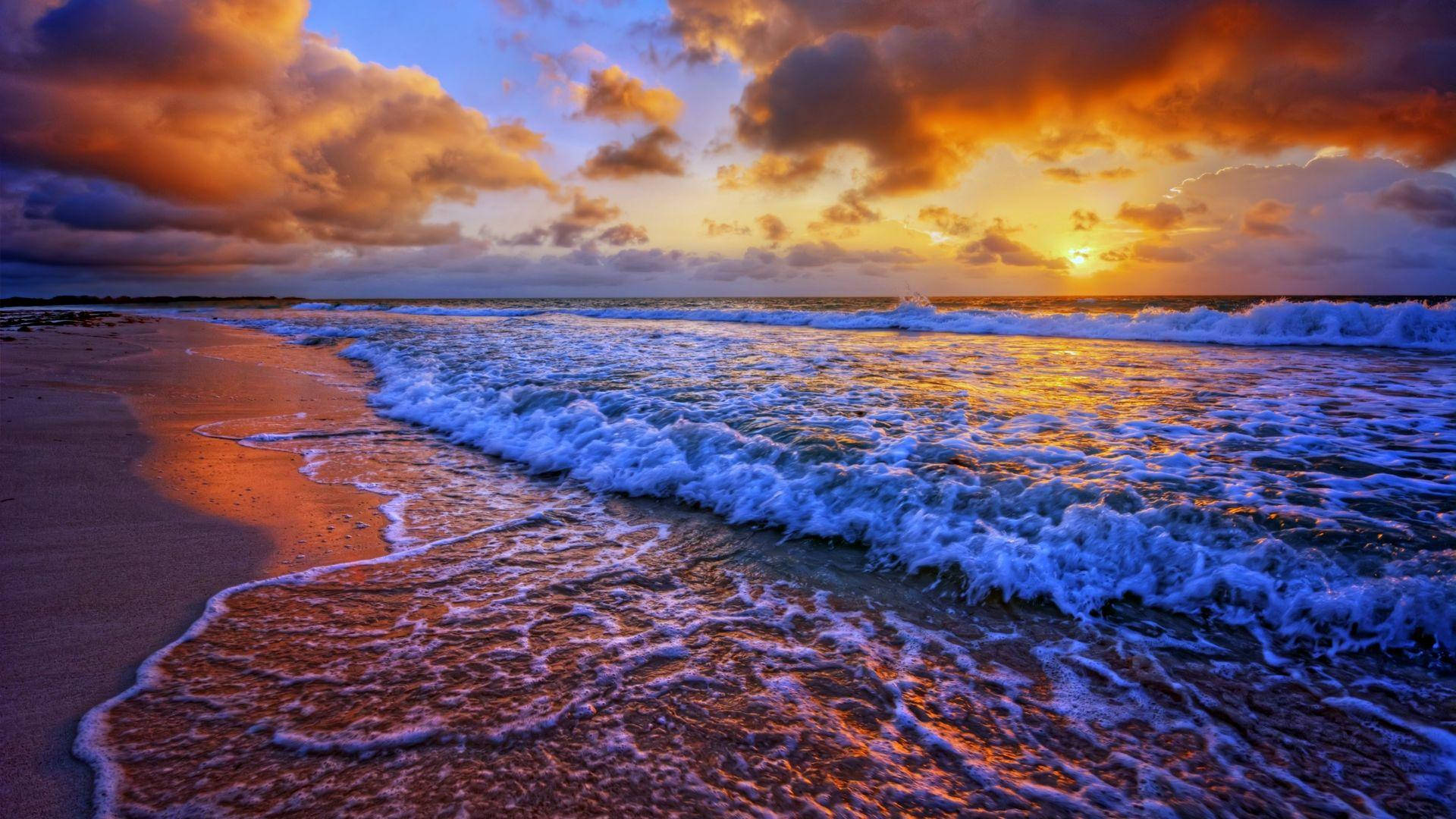 暗色日落時的海灘邊浪花 壁紙 — 夕陽將海浪和沙灘染成火熱的橙色和深藍色調。
