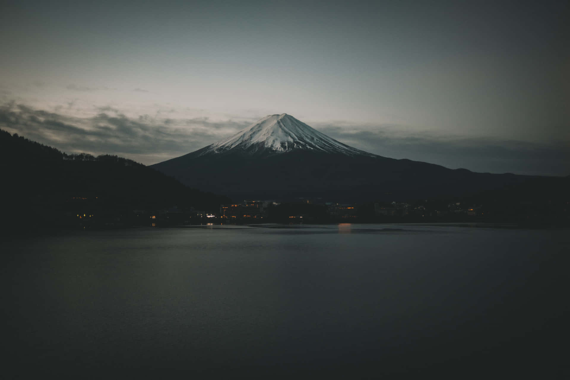 黃昏時分的富士山倒映在平靜的湖面上，遠處有城鎮燈光。