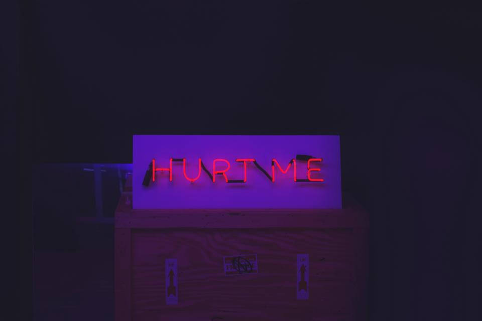 暗色霓虹 iPhone「傷我」 壁紙 — 一個發光的紅色霓虹燈牌，上面寫著「HURT ME」，在黑暗的房間裡將紫色光暈投射在木箱上。