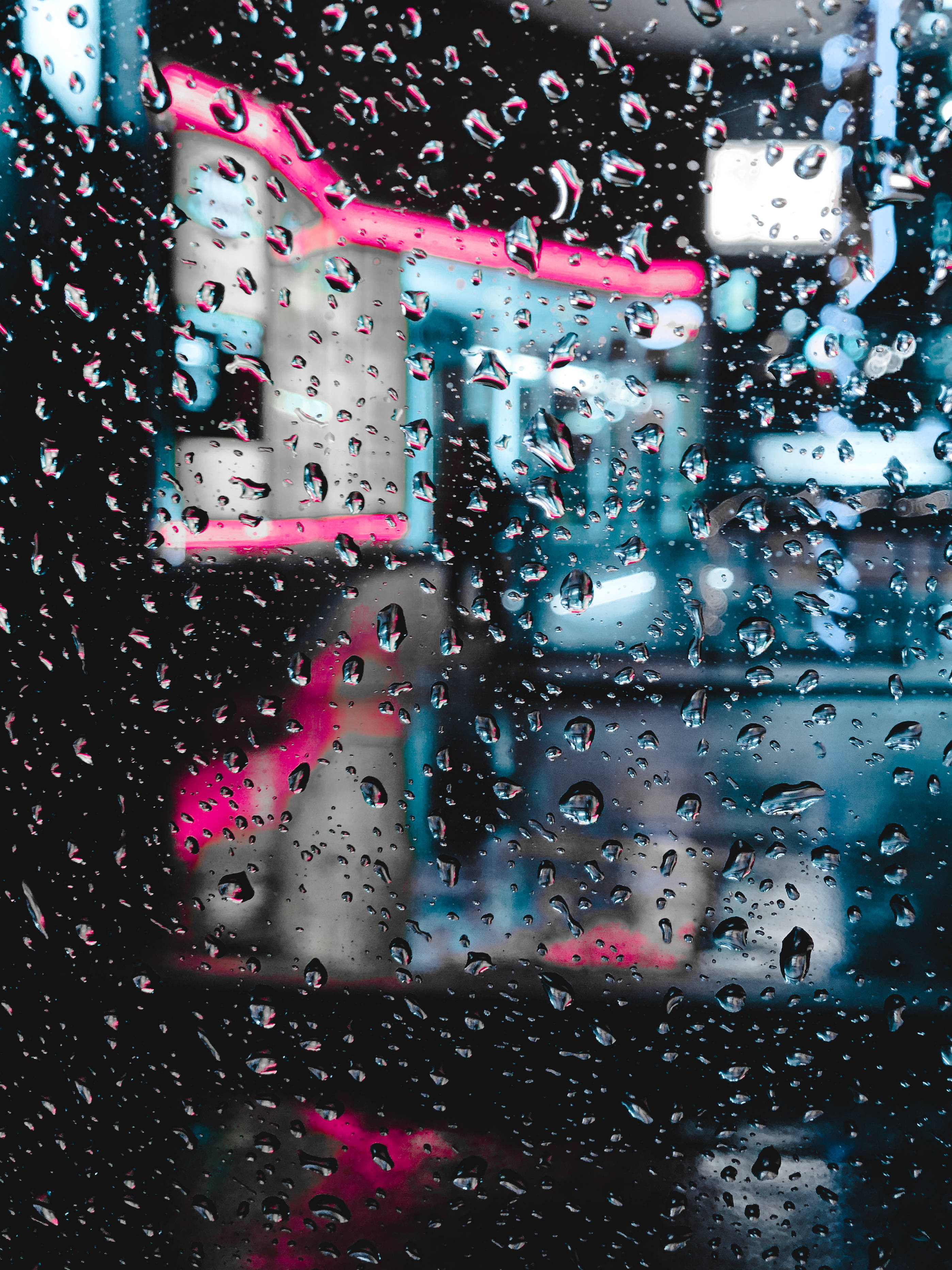暗色霓虹 iPhone 玻璃上的雨滴 壁紙 — 雨滴沿窗戶流下，顯露出夜間城市模糊的霓虹燈光。