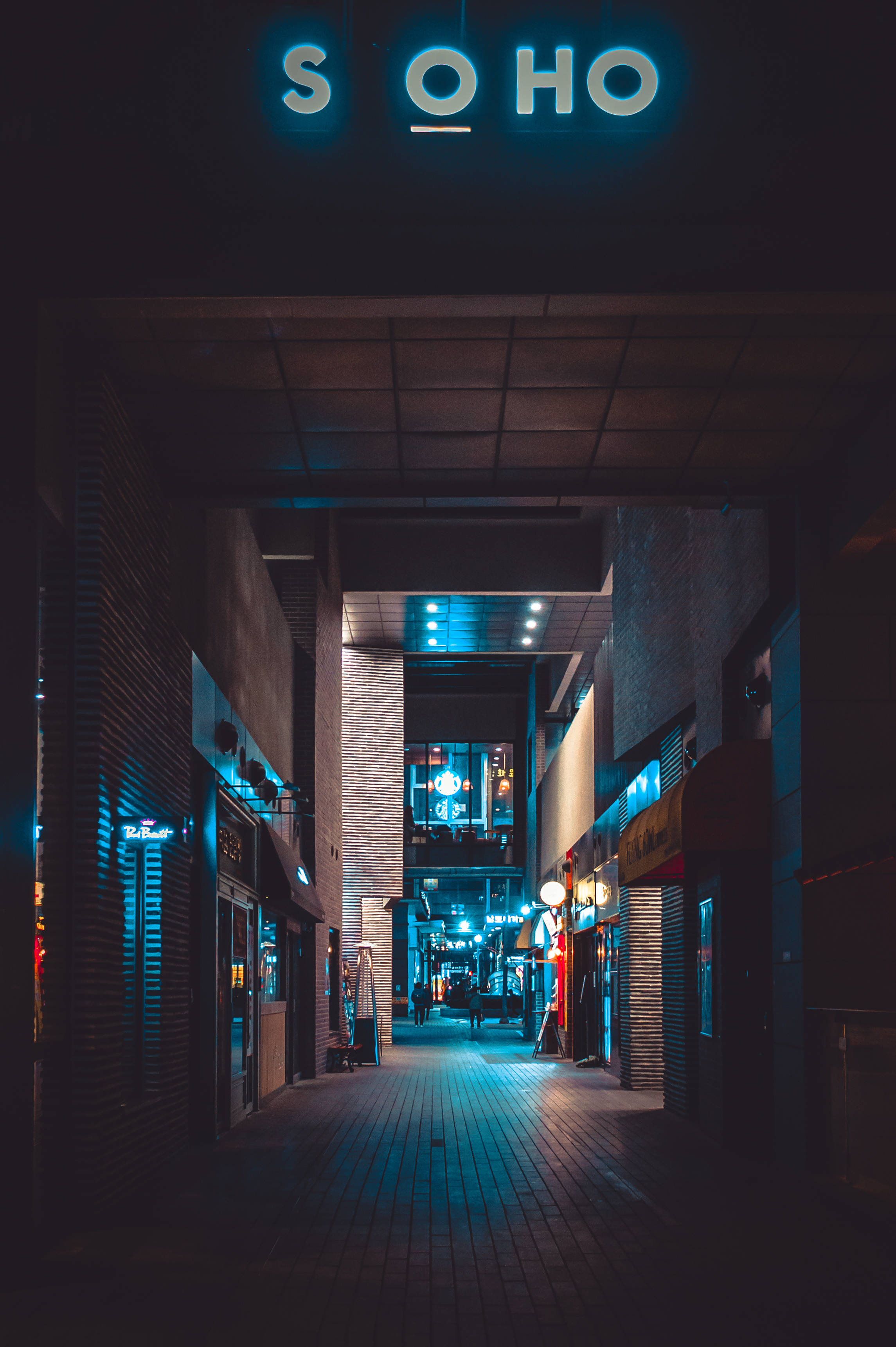 暗色霓虹 iPhone 苏豪區招牌 壁紙 — 夜晚狹窄的城市小巷，被發光的藍色霓虹燈招牌和溫暖的路燈照亮，投下長長的影子。