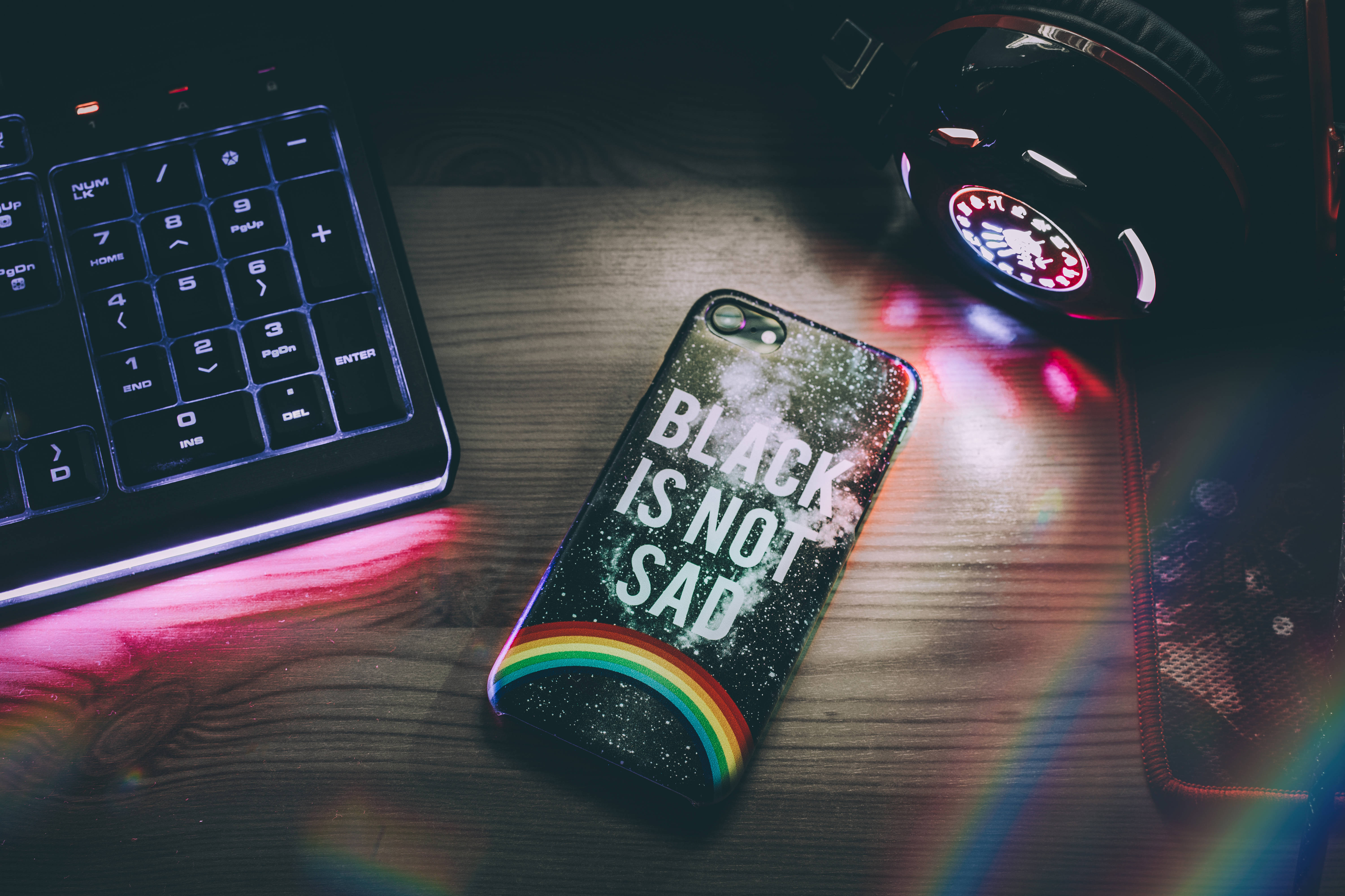 暗色霓虹 iPhone 靠近鍵盤 壁紙 — 一個閃閃發光的手機殼，帶有彩虹和文字，放在木桌上，旁邊有鍵盤和耳機，在色彩繽紛的光暈下。