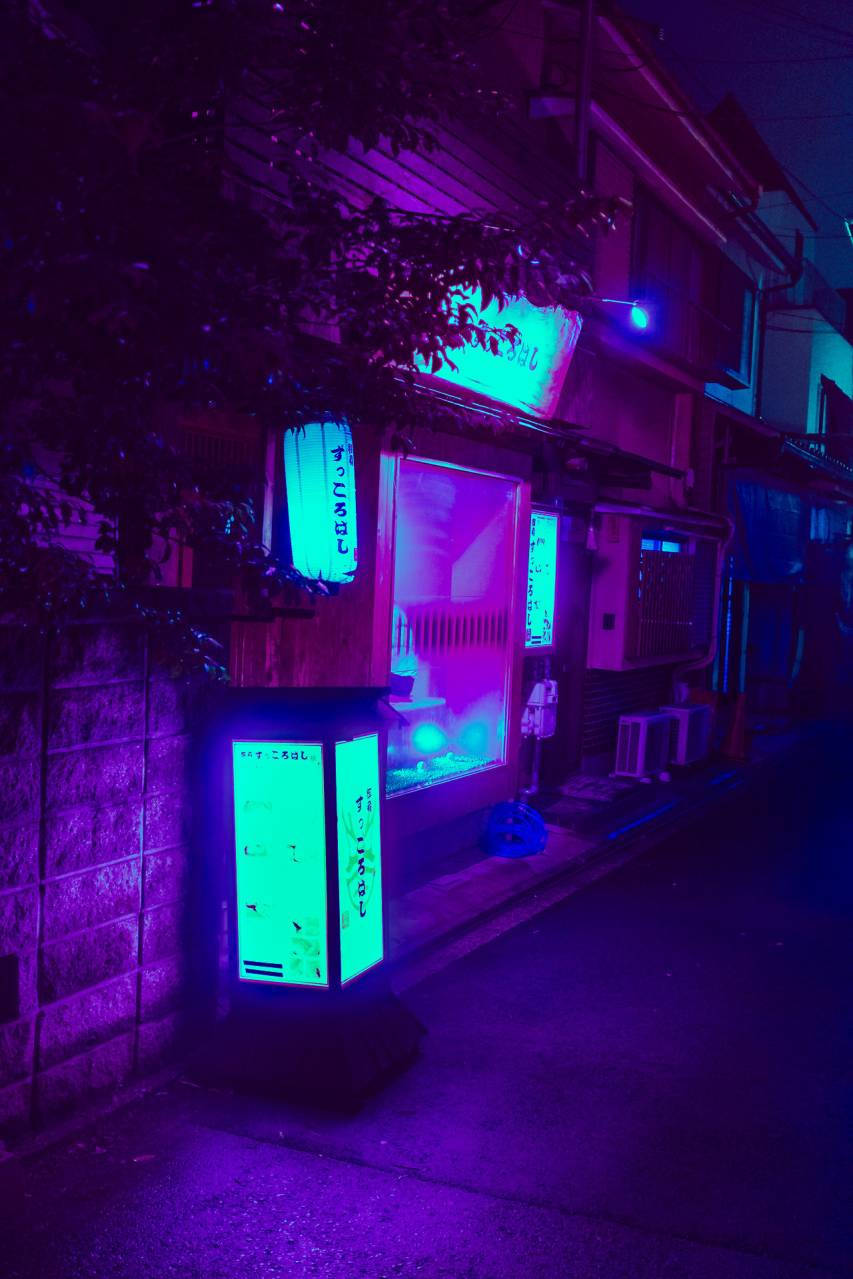 暗色霓虹日本攤位iPhone壁紙 壁紙 — 夜晚一條霓虹燈照亮的日本小巷，綠色發光的自動售賣機和紫色環境光在石牆和樹木上投下陰影。