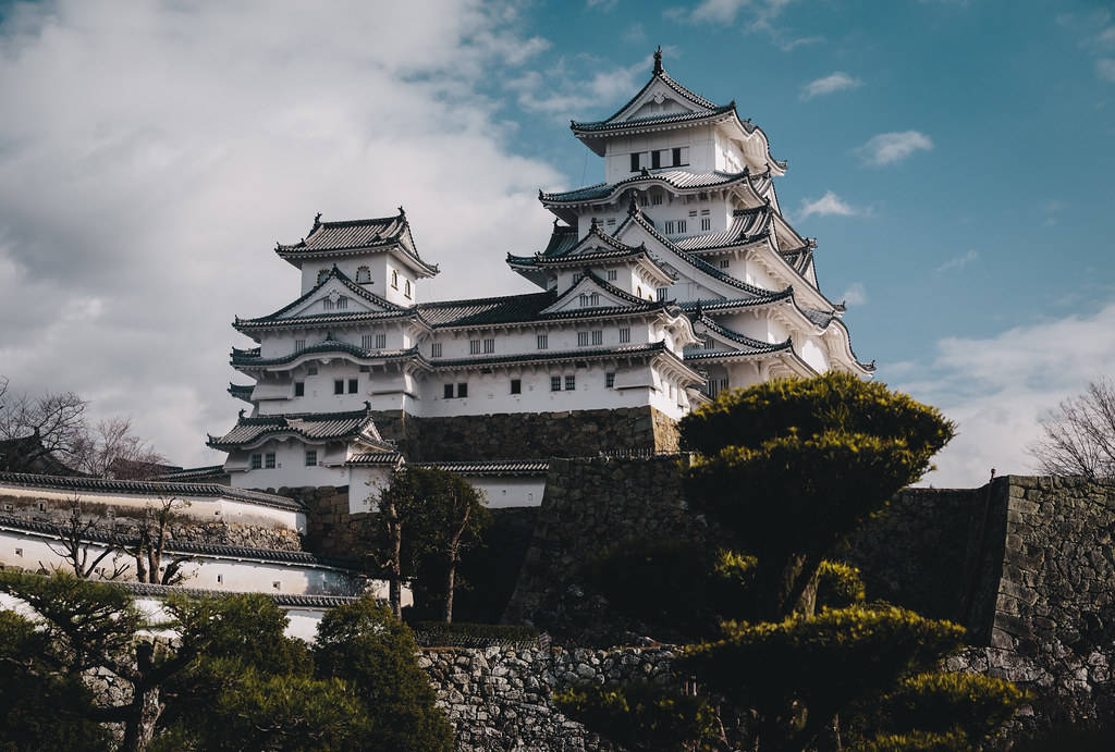 白色日本城堡在石山上，周圍有綠樹和部分多雲的天空