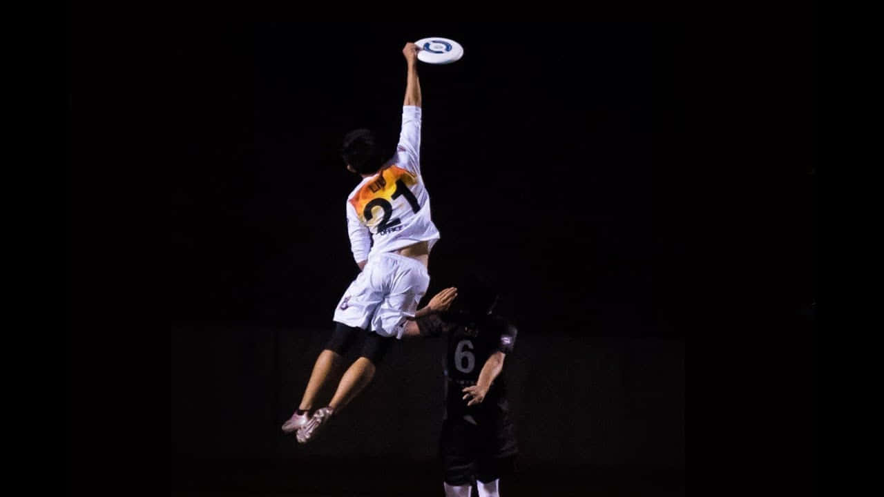 暗黑風格最佳 Ultimate Frisbee 背景 壁紙 — 一名白衣球員高高跳起接飛盤，另一名黑衣球員在下方防守，背景昏暗。