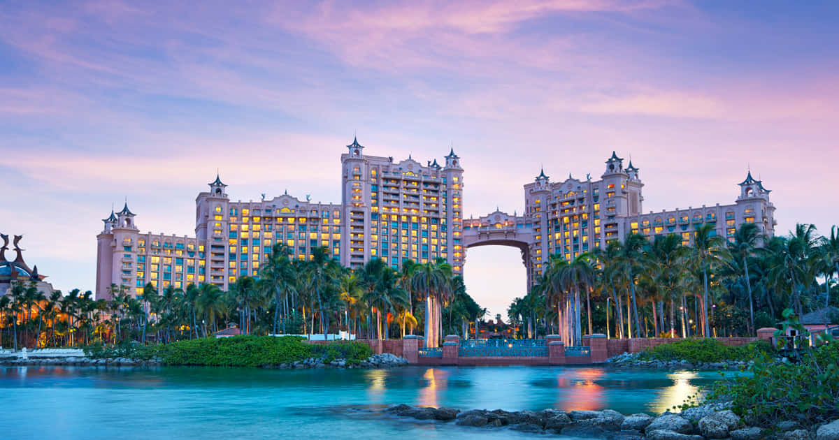 Bahamas Island 桌布 壁紙 — 暮色中，大型粉紅色度假酒店，窗戶閃爍，棕櫚樹倒映在碧綠的水面上。