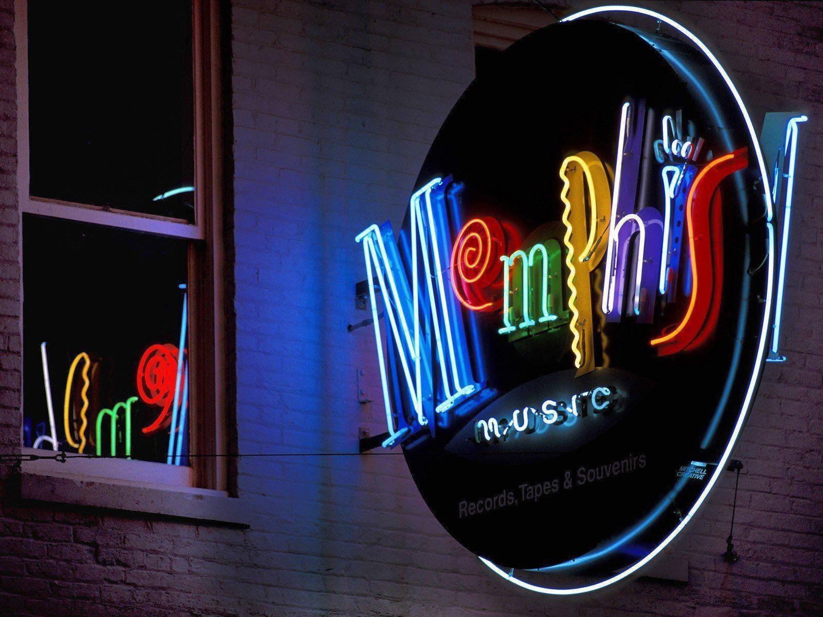 暮色中充滿活力的孟菲斯市中心 壁紙 — 一個寫著「Memphis Music」的鮮豔霓虹招牌在夜間映照著磚牆，並在窗戶上投射出色彩繽紛的倒影。