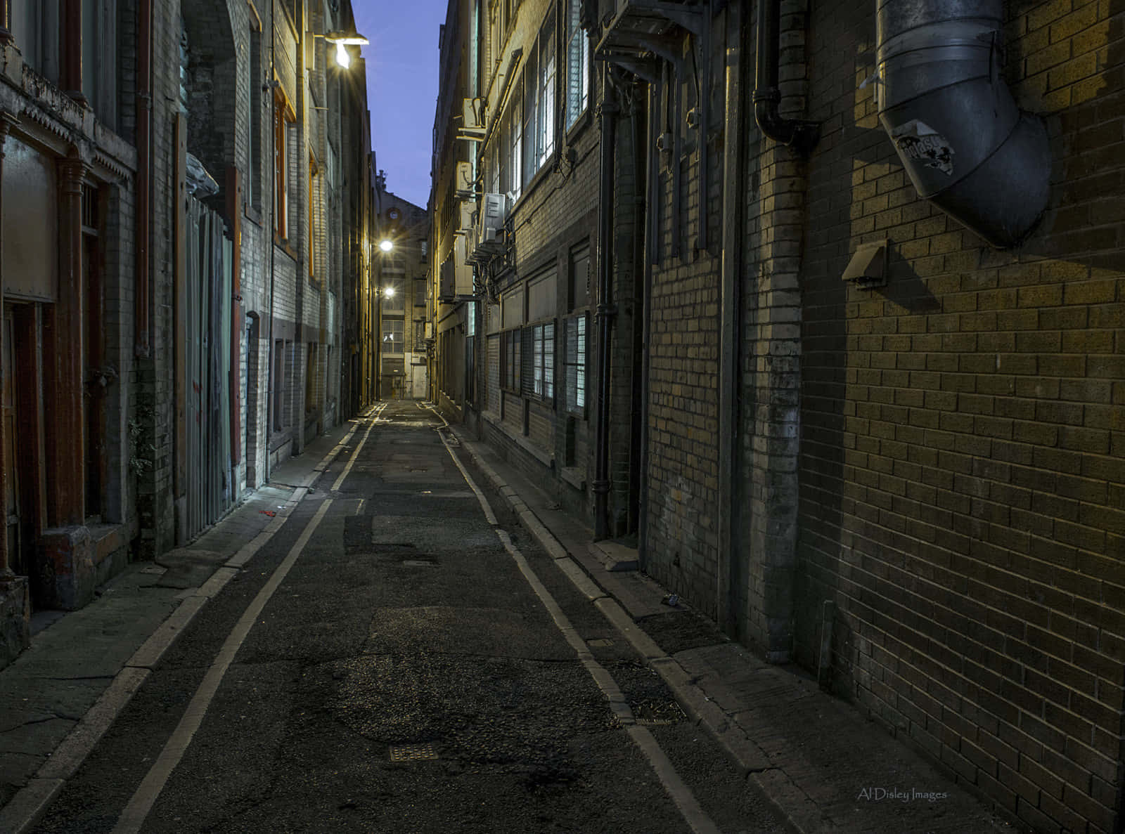 Alleyway 桌布 壁紙 — 暮色中狹窄的磚砌小巷，溫暖的路燈在潮濕的路面上投下長長的影子。