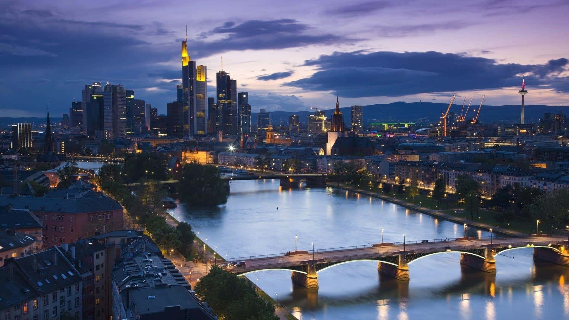 Germany 桌布 壁紙 — 暮色中的城市景觀，河流流經燈火通明的摩天大樓和橋樑，天空呈紫色。