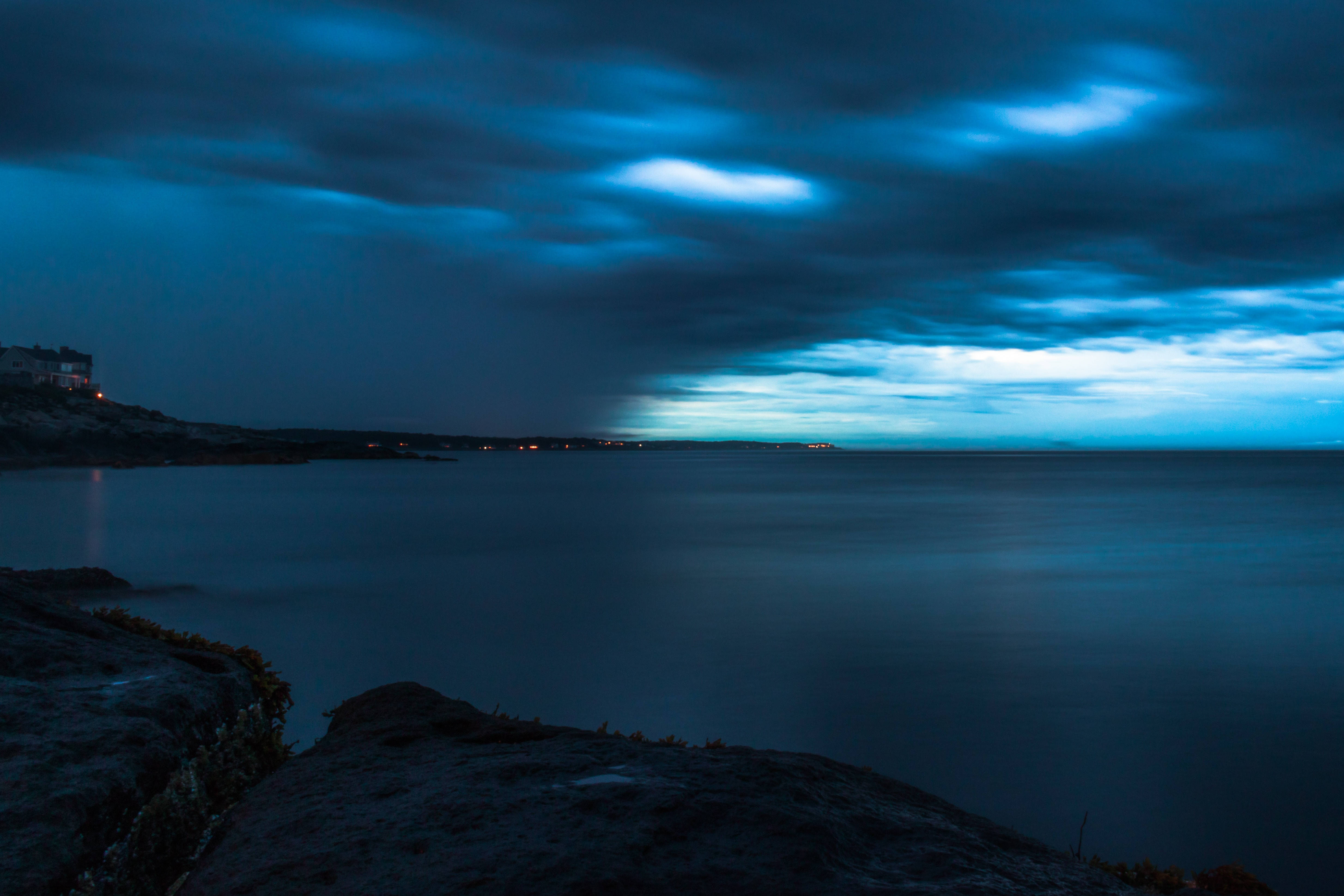 暮色中的暴風雲天際 壁紙 — 寧靜的黃昏海景，發光的藍色雲朵在平靜的水面上，遠處有海岸燈光。
