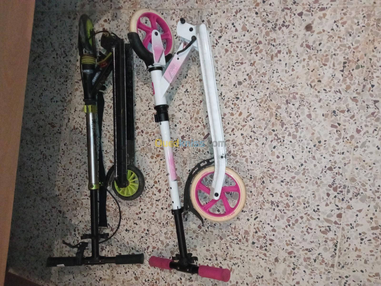 暮色中的神祕山景 壁紙 — 兩部摺疊滑板車，一部黑色配綠色車輪，另一部白色配粉紅色車輪，靠在一片斑點水磨石地板上。