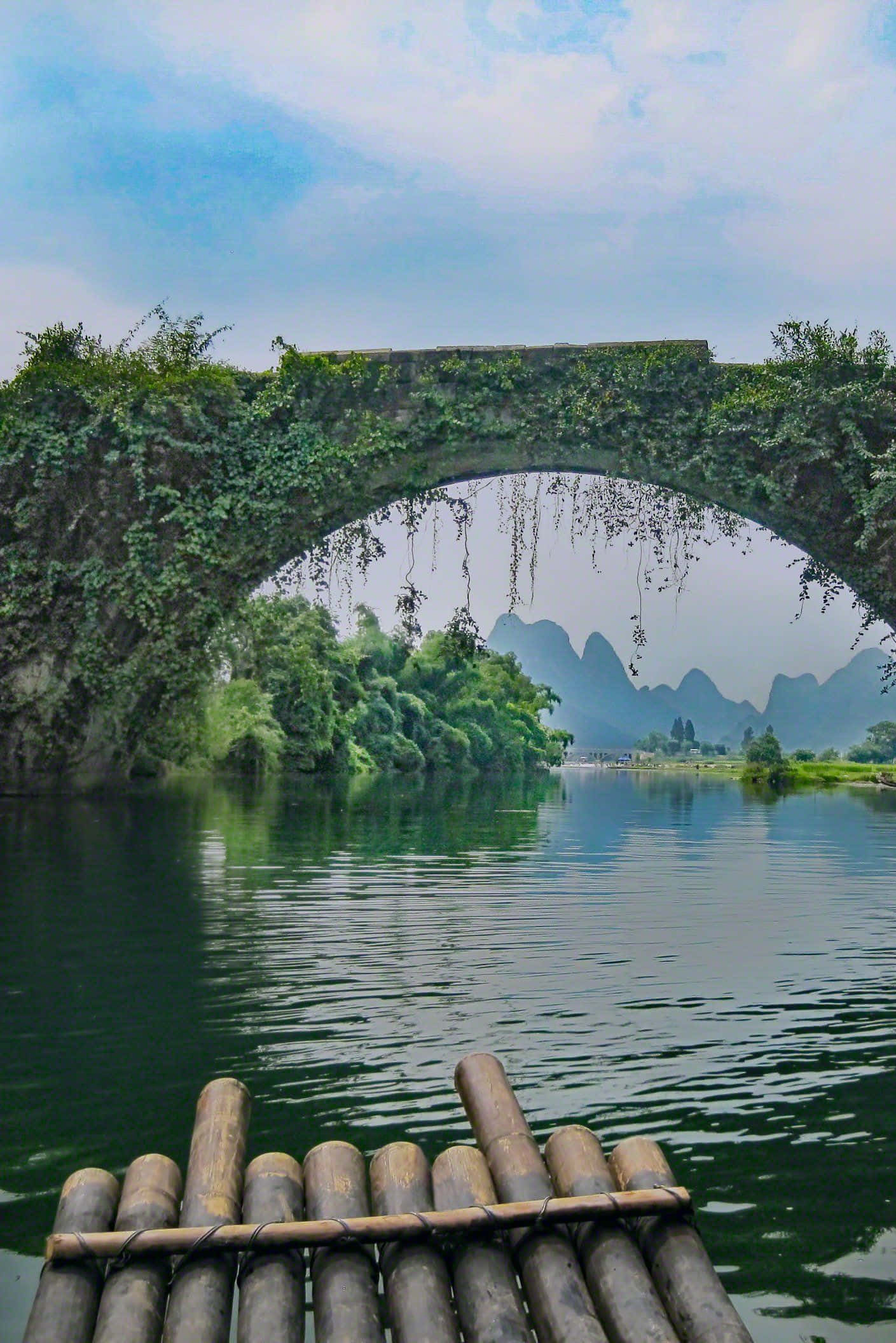 暮色橋影倒映河面 壁紙 — 竹筏漂浮在長滿青苔的石拱橋下，倒映在平靜的綠水中，遠處是喀斯特地貌的山巒。