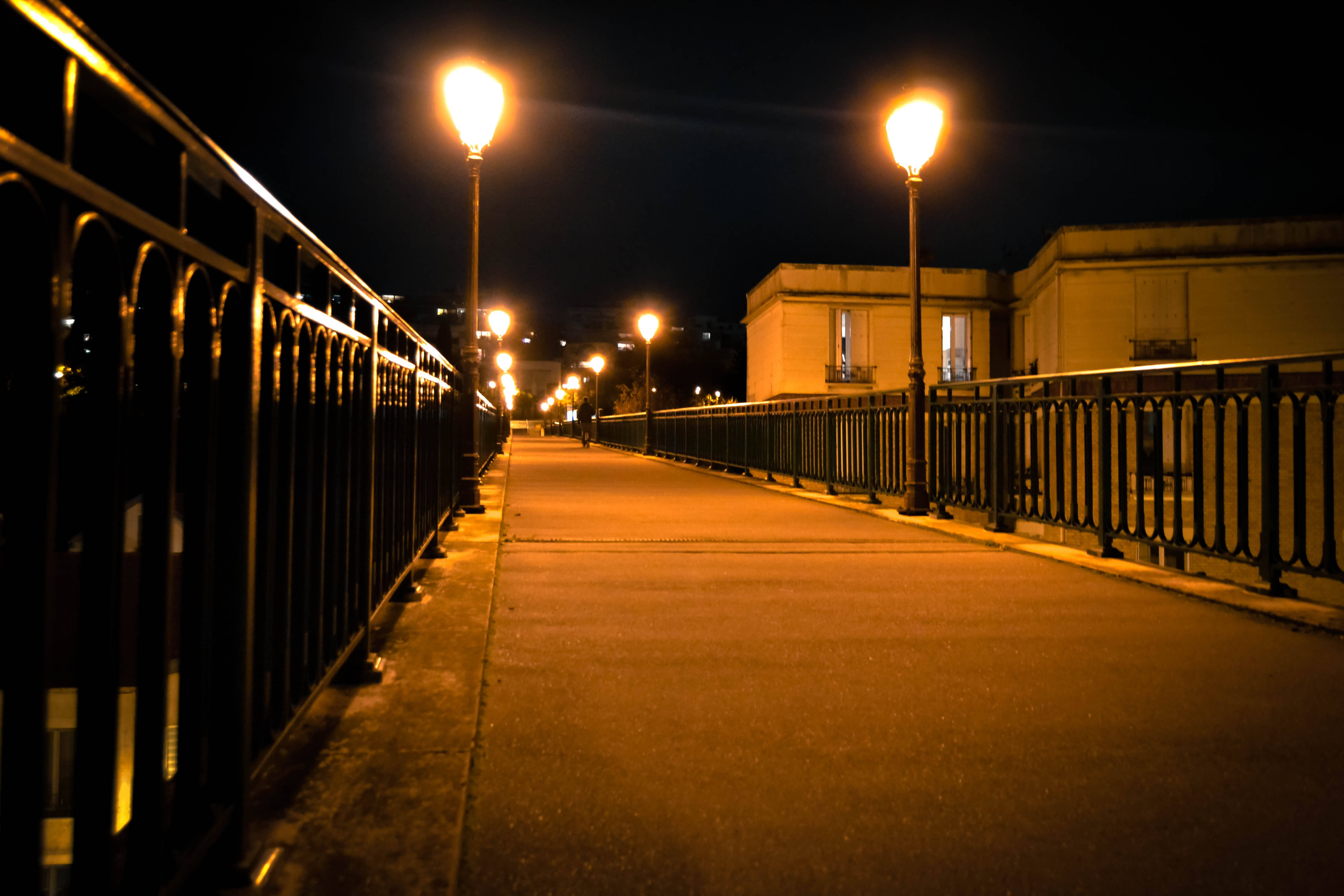 暮色橙城街景，燈光溫柔點綴 壁紙 — 一條寧靜的夜間行人道，由溫暖發光的街燈照亮，在鋪砌的小路上投下長長的影子，兩旁是華麗的欄杆。