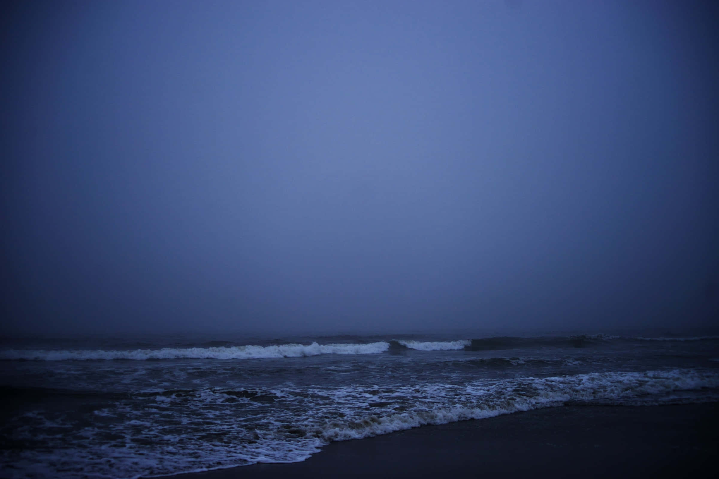 Dark Ocean 桌布 壁紙 — 暮色蒼茫的海灘場景，深藍色的波浪在朦朧的天空下拍打著霧濛濛的海岸。