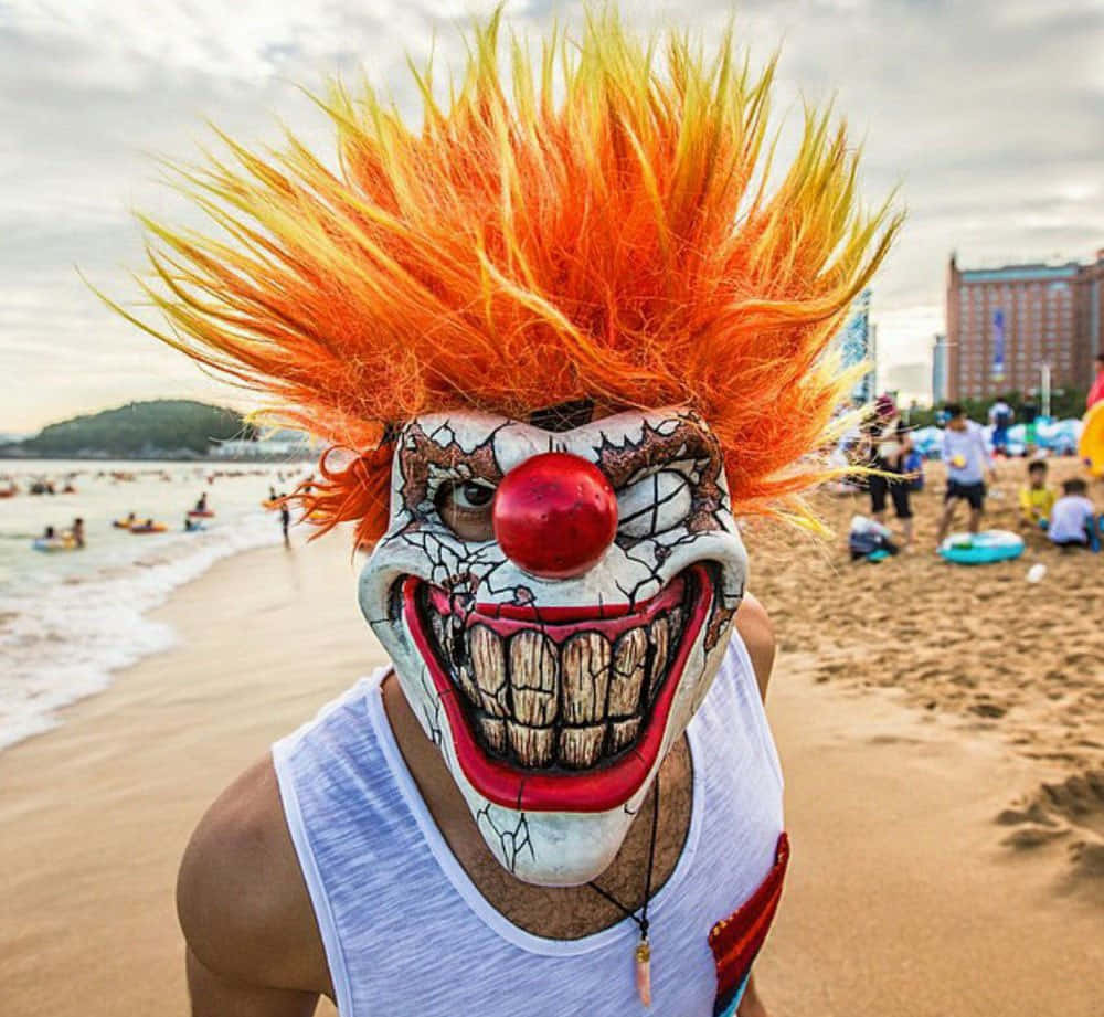 暴躁搞笑小丑圖 壁紙 — 一個戴著怪誕小丑面具和火橙色假髮的人站在擁擠的海灘上，海浪在附近拍打。