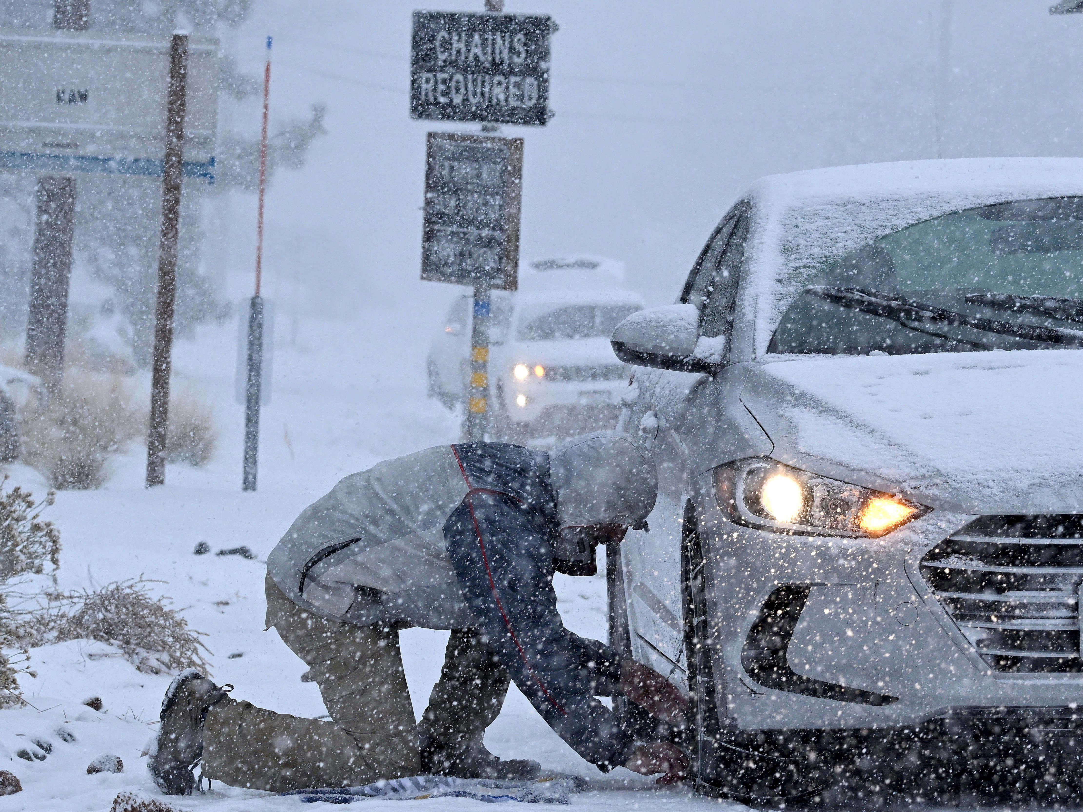 暴雪可將你從春日暖意帶入冬日仙境 壁紙 — 一個人跪在厚雪中，在「必須使用鏈條」標誌下為白色汽車安裝輪胎鏈。