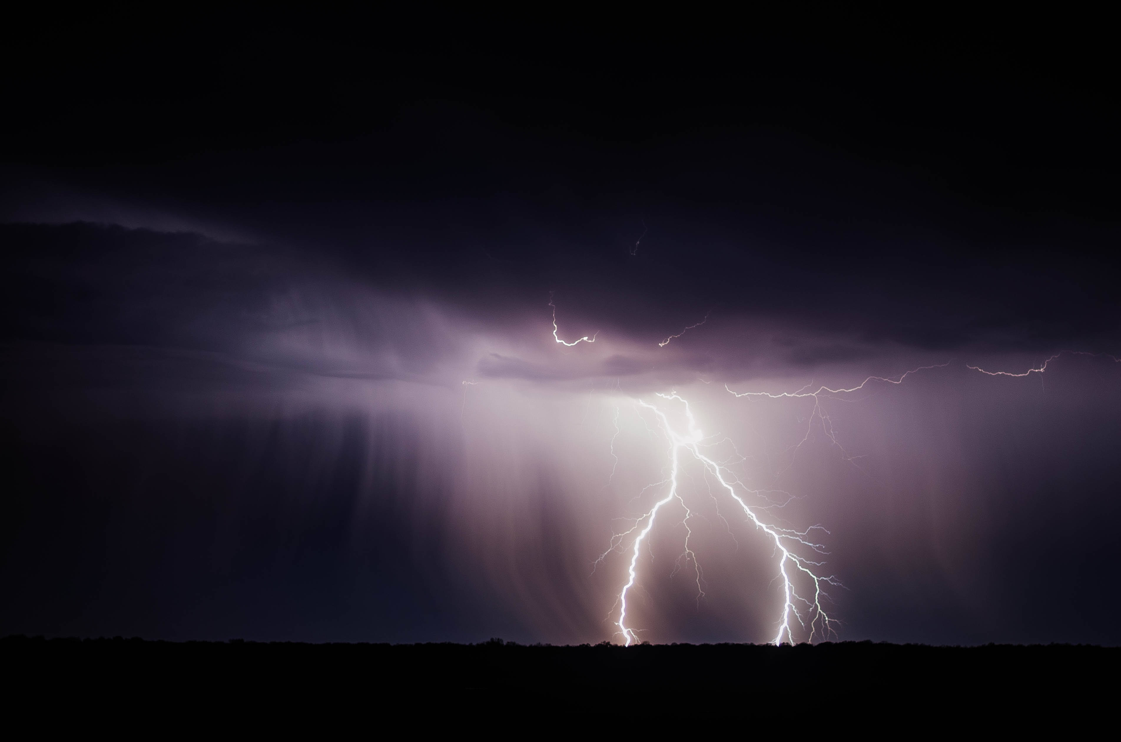暴風雨中的閃電 壁紙 — 一道強大的閃電劃破黑暗的暴風雨天空，映襯著剪影般的地平線，雨水傾瀉而下。