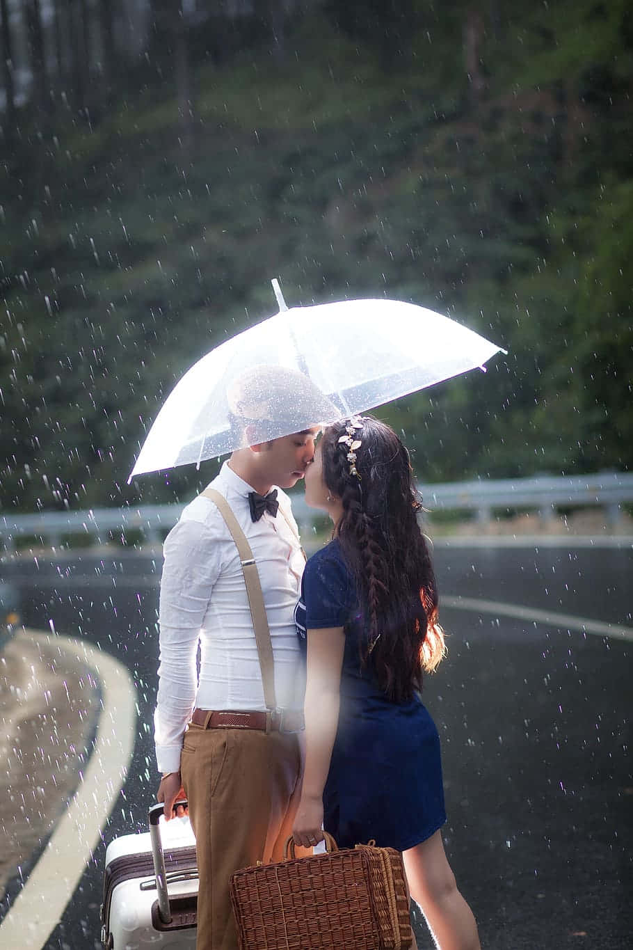 暴風雨中相擁的戀人 壁紙 — 一對情侶在雨中的路邊共享透明雨傘和溫柔的親吻，周圍是茂密的綠樹，並手持復古行李箱。