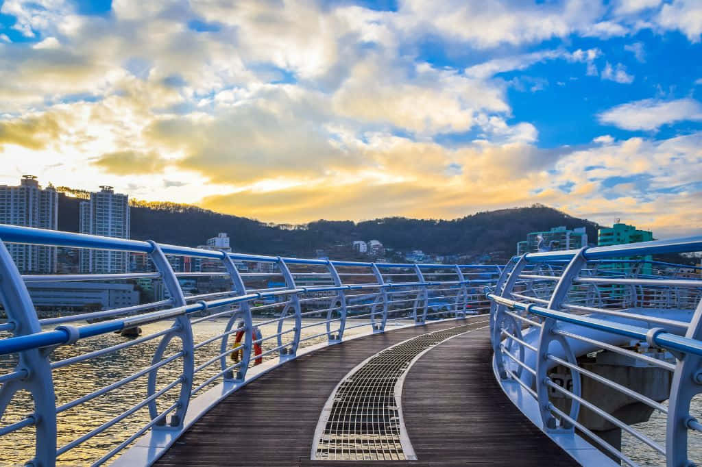 日落時分，帶藍色欄杆的彎曲木橋橫跨水面，背景有城市建築。