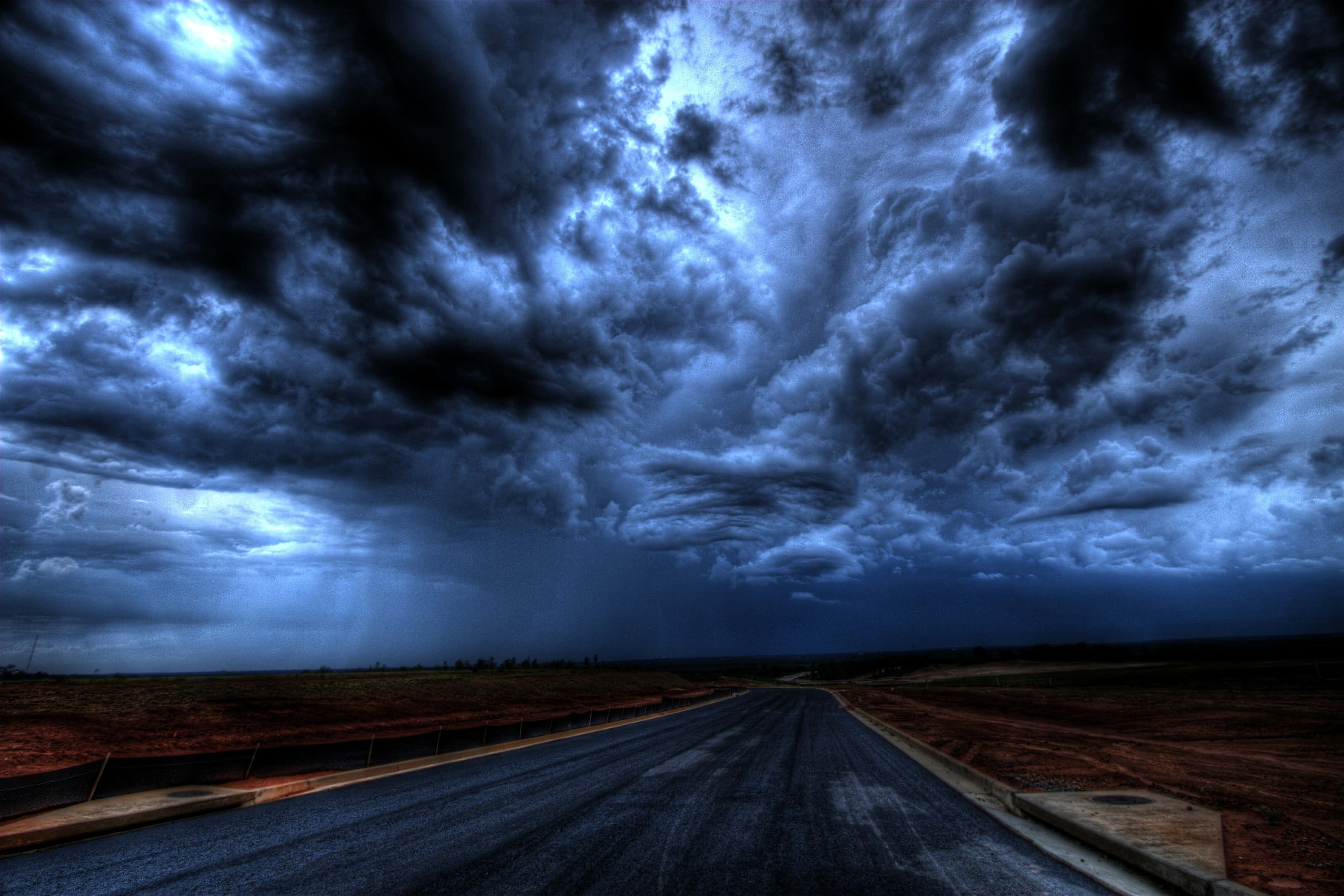 暴風雲籠罩的黑暗天空與道路 壁紙 — 一條孤獨的瀝青路延伸至暴風雨的地平線，天空佈滿戲劇性的藍色和黑色雲層。