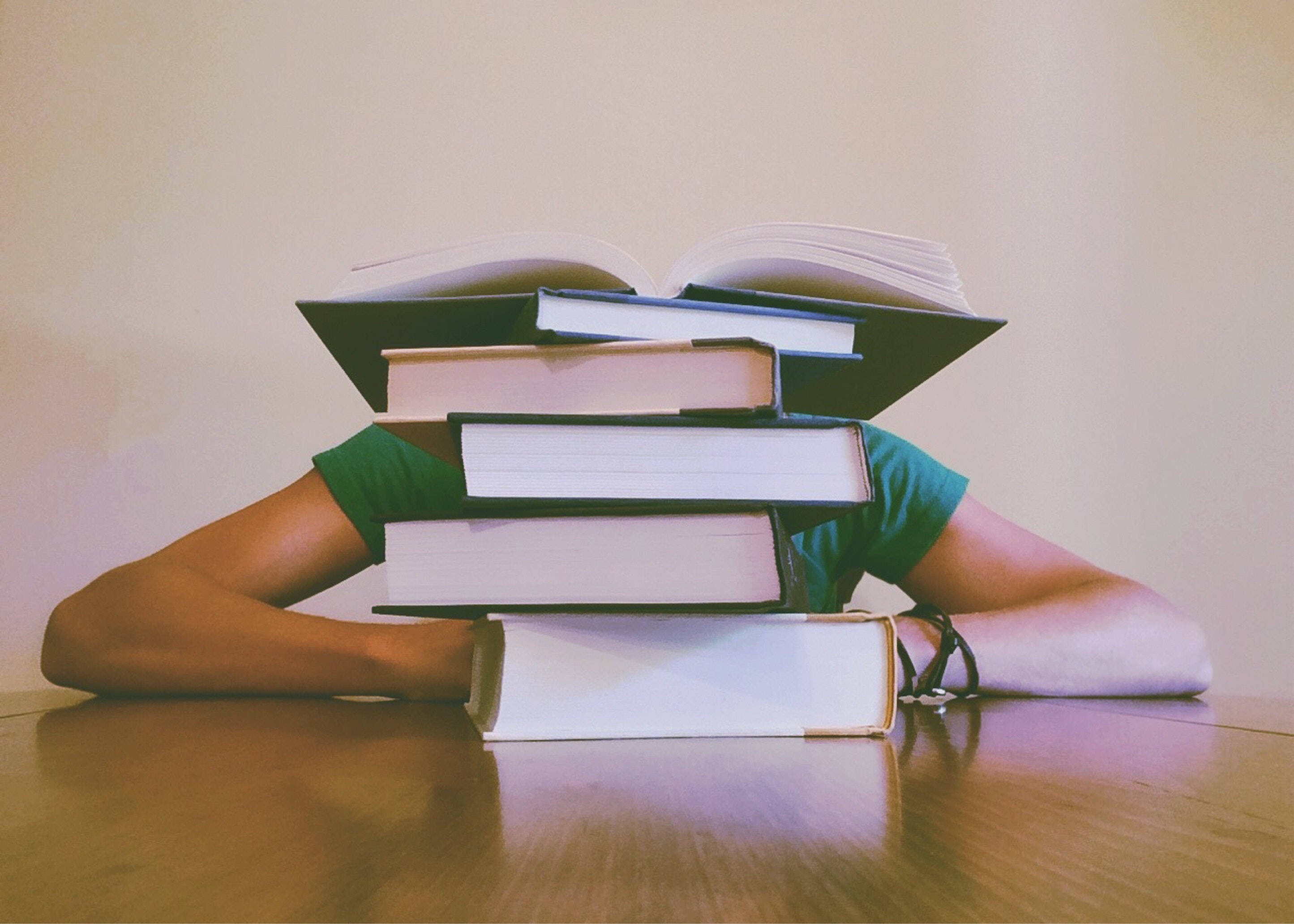 書堆中的學習 壁紙 — 一個穿著綠色襯衫的人將頭靠在木桌上的一疊高高的書本下。