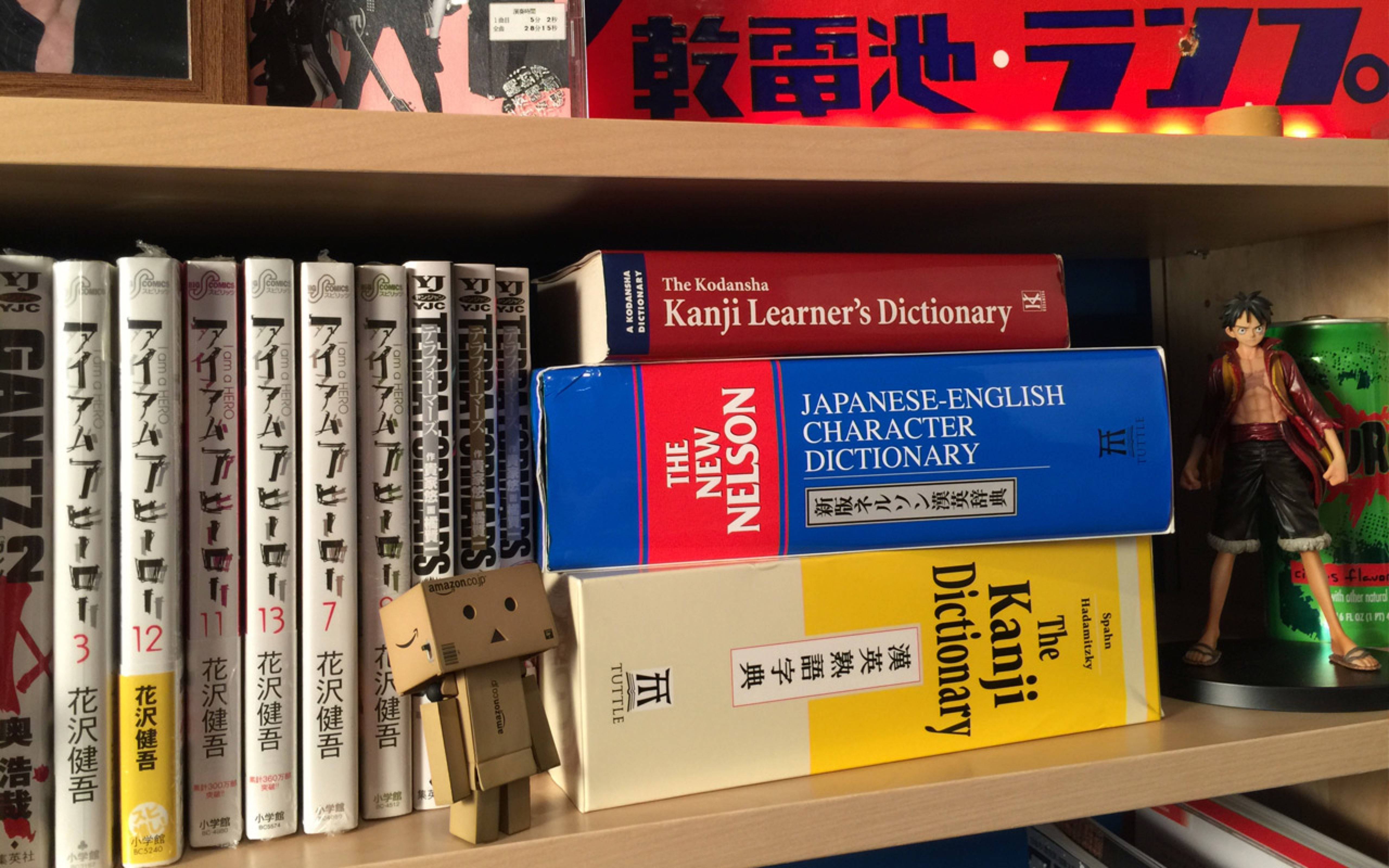 書架上的字典 壁紙 — 木書架上展示日本漫畫卷、字典和動漫人偶，在溫暖燈光下。