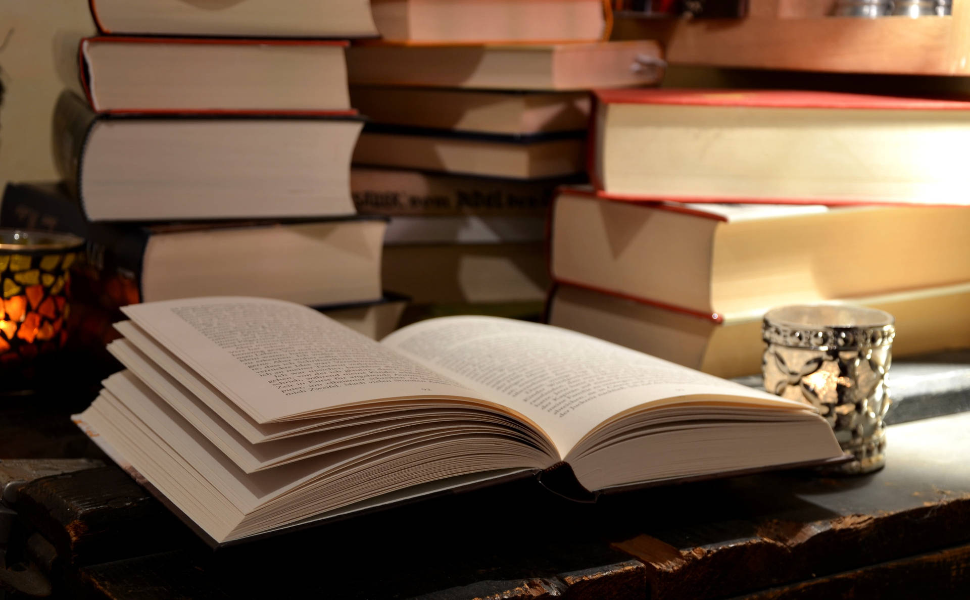 木桌上的打開的書，附近有成堆的書籍和點燃的蠟燭。