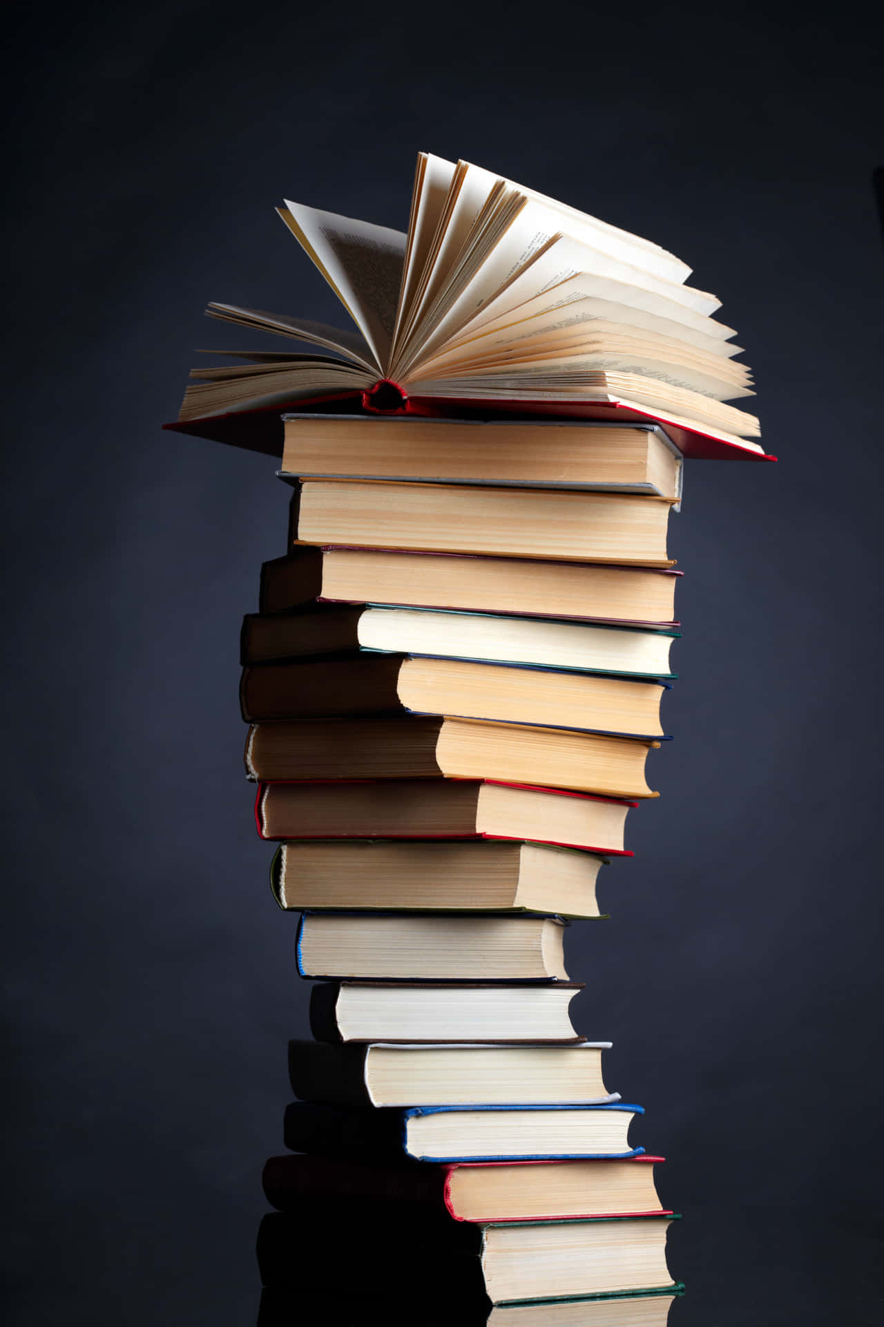 高聳的書堆，頂部有一本打開的書，置於深色反光表面上。