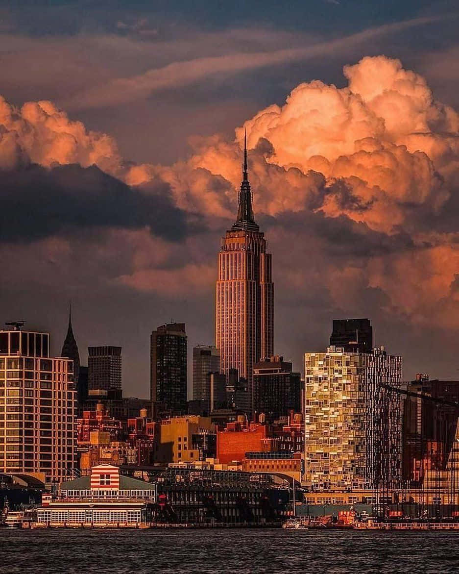 曼哈頓夜景燈光璀璨 壁紙 — 帝國大廈在日落時分聳立於紐約天際線之上，上方有火紅的橙色雲彩，下方有水。