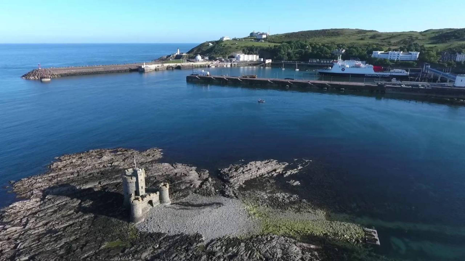 曼島海港碼頭風景 壁紙 — 歷史石城堡嘅航拍圖，位於岩石海岸，旁邊係有船同綠色山丘嘅港口，天空晴朗蔚藍。
