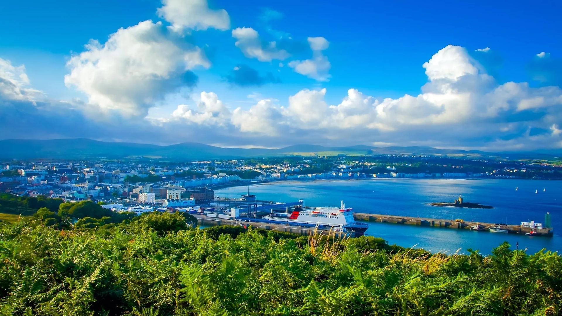 曼島港口的絕美風景 壁紙 — 一個充滿活力的沿海小鎮，坐落在深藍色的港灣旁，一艘大型渡輪停泊在晴朗的天空下，天空佈滿蓬鬆的白雲。