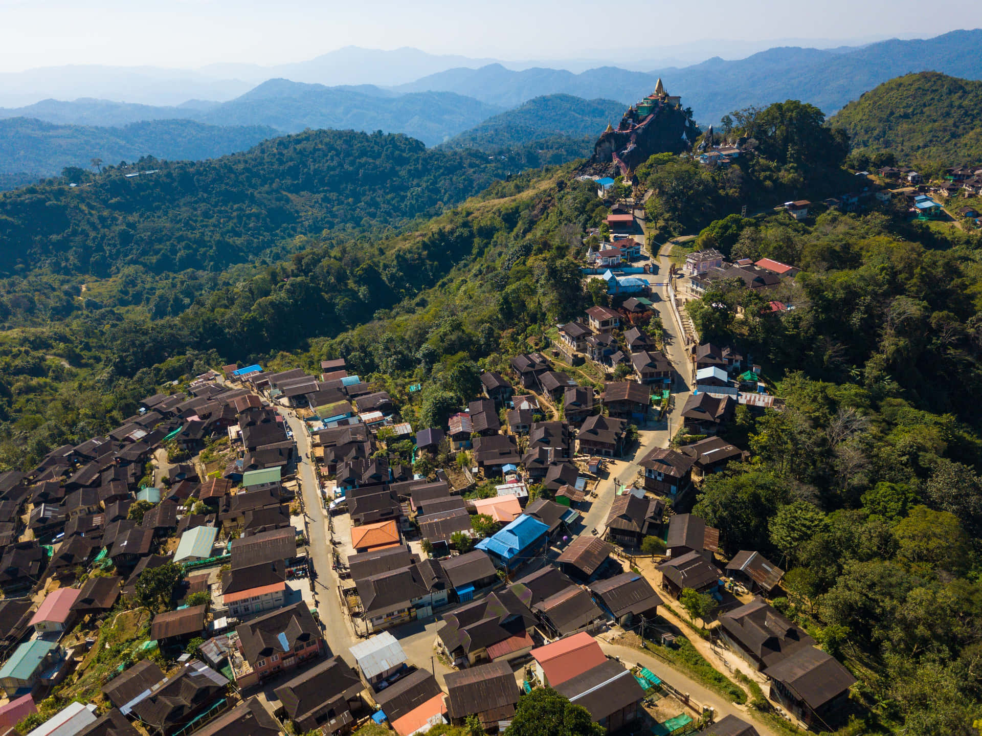 山村，房屋沿著林木茂密的山坡向上攀爬，通往山頂的一座寺廟。